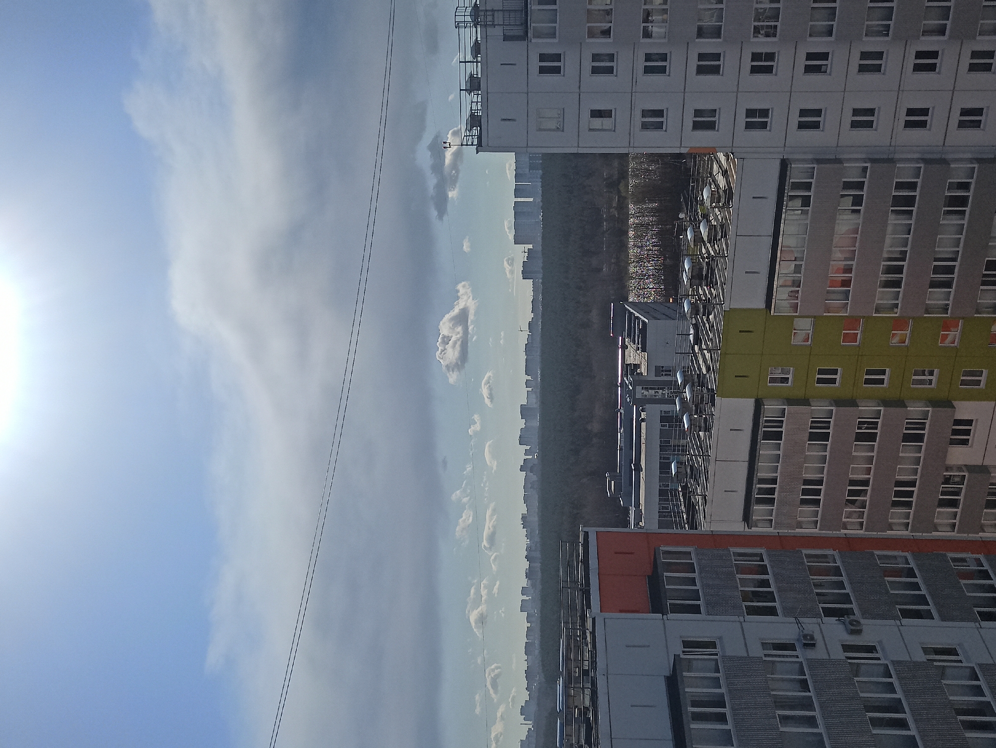 This image captures an urban landscape viewed from a high vantage point, possibly an upper floor of a building. The scene is dominated by multi-story residential or commercial buildings under a partly cloudy sky.
In the foreground and mid-ground, several large, block-like buildings with numerous windows are prominent. Their facades vary, with some being light grey or white, others a muted yellow-green, and one featuring a distinct orange-red section. Many windows are visible across these structures, with some displaying reflections. Rooftop structures, possibly ventilation systems or maintenance units, are seen on several buildings, along with what appear to be satellite dishes or similar equipment on one of the mid-ground buildings.
The sky occupies the upper portion of the image. The left side features a bright, sun-drenched blue sky with a visible sun flare, indicating bright daytime. The right side is covered by a large, diffuse cloud formation. Further into the distance, a band of smaller, puffier clouds is scattered across the horizon.
Below the horizon line of the buildings, a dense stretch of trees or a forest extends across the mid-distance. Beyond this green belt, a distant, hazy skyline of numerous other high-rise buildings can be seen, indicating a vast urban sprawl. Two dark, thin wires or power lines stretch diagonally across the top-left portion of the sky.
The weather appears to be bright and clear in parts, with significant cloud cover elsewhere. There are no people visible, and no discernible text is present in the image.