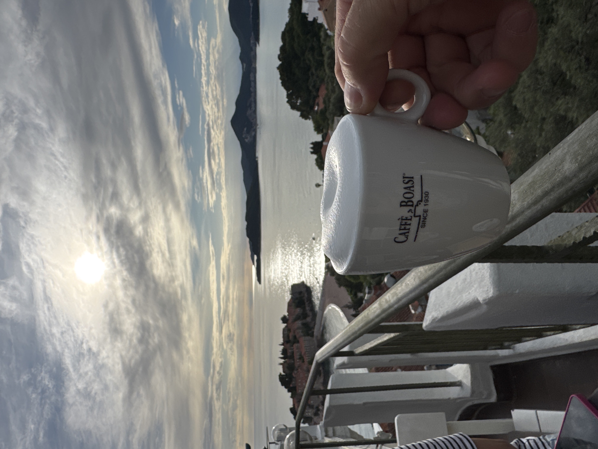 Here's a detailed description of the image:
**Overall Scene:**
The image presents a partial view of a scene overlooking a coastline and the sea. The composition is dominated by a coffee cup held by a person, with the landscape serving as a backdrop.
**Location:**
Likely Sveti Stefan, Montenegro, based on the coastal scenery with islands or peninsulas visible.
**Foreground:**
* **Person:** Only a hand holding a white coffee cup is visible.
* **Object:** A white coffee cup with the logo "Caffe BOASI since 1930" is the central object.
* **Railing:** A railing with white supports and a gray handrail is present.
**Background:**
* **Sea:** A body of water (the sea) stretches into the distance, reflecting sunlight.
* **Coastline:** Islands or peninsulas are visible along the coastline, covered with vegetation.
* **Sky:** The sky is filled with clouds, with the sun providing illumination.
* **City:** Part of a city with buildings and vegetation can be seen on a hill.
