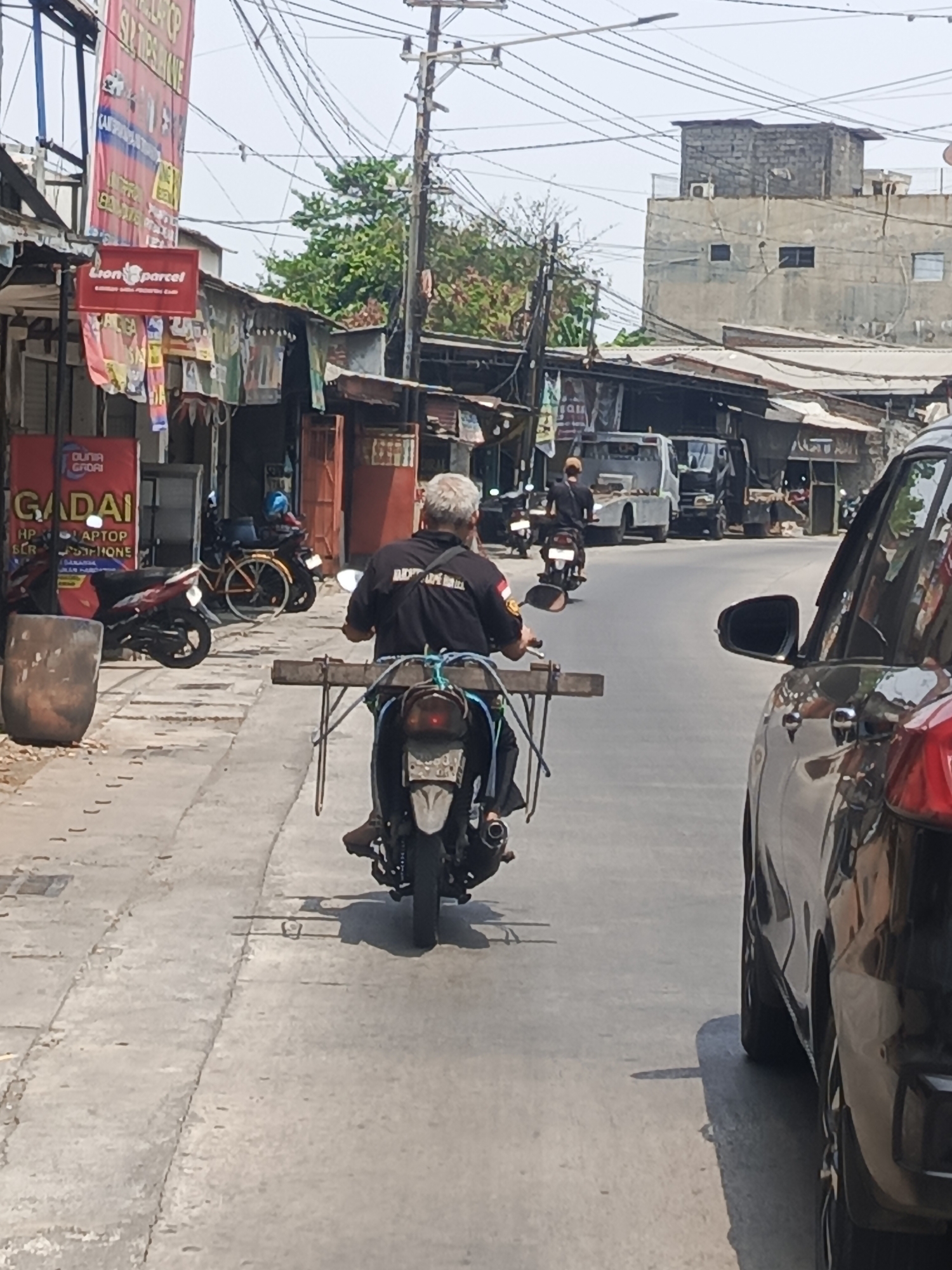 In the city of Kepuh Kiriman, Indonesia, a man with graying hair is riding a motorcycle on a paved road. He is wearing a black t-shirt and dark pants and is carrying a long wooden plank horizontally across the back of his motorcycle, secured with ropes. The motorcycle has a license plate that appears to be from Indonesia. The man is positioned in the middle of the road, with a black car partially visible in the right foreground and a variety of buildings and shops lining the left side of the road. These buildings have corrugated metal roofs and some display advertising banners, including one for "Lion Parcel" and another for "DUNIA GADAI". There are other motorcycles and a small truck in the background, suggesting a busy street scene. The sky is bright and clear.