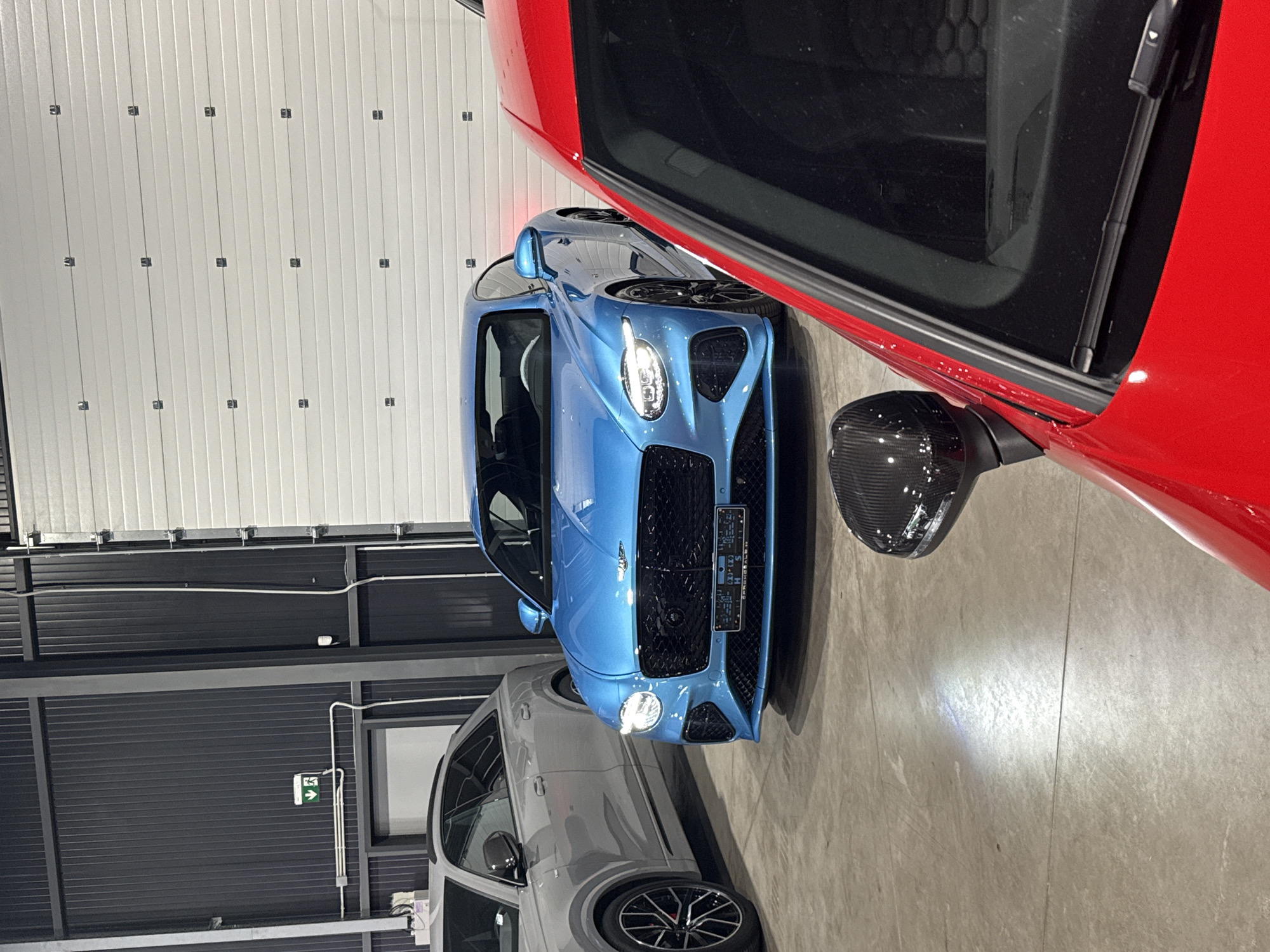 The image captures a detailed interior scene featuring multiple luxury cars. In the foreground, the hood and windshield of a vibrant red sports car are prominently displayed, with a carbon fiber side mirror visible to the left. Behind it, a striking blue Bentley Continental GT is parked, its distinctive grille and headlights illuminated. To the left of the Bentley, a silver Audi SUV is partially visible. The cars are situated on a concrete floor within a modern industrial-looking garage or showroom, characterized by corrugated metal walls and a large, segmented white overhead door in the background. The environment appears to be clean and well-lit, suggesting a showroom or a private garage for high-end vehicles. There are no people visible in the frame. This scene is located in Plovdiv, Bulgaria, specifically within the premises of "Premium Auto".