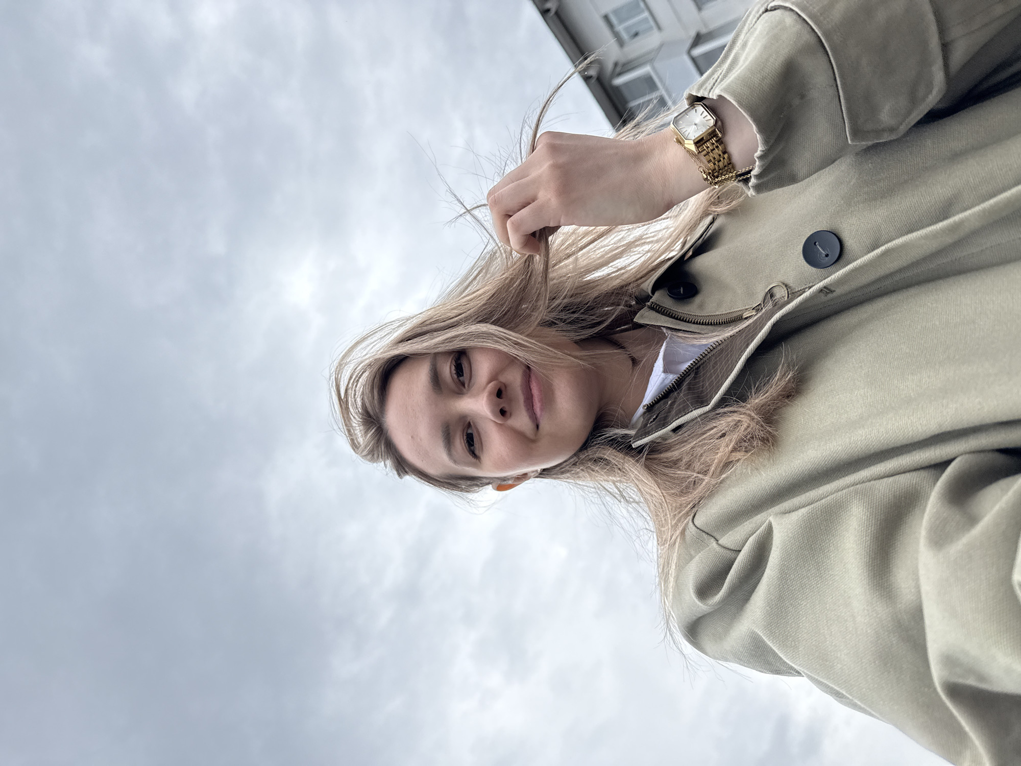 A low-angle shot captures a woman outdoors. The overcast sky, indicative of cloudy weather, serves as the background. She has blonde hair and is wearing a light green trench coat. A gold watch is visible on her wrist. She is holding a strand of her hair. A building is partially visible in the upper right corner. The location may be the city of Chaikovsky, Russia.