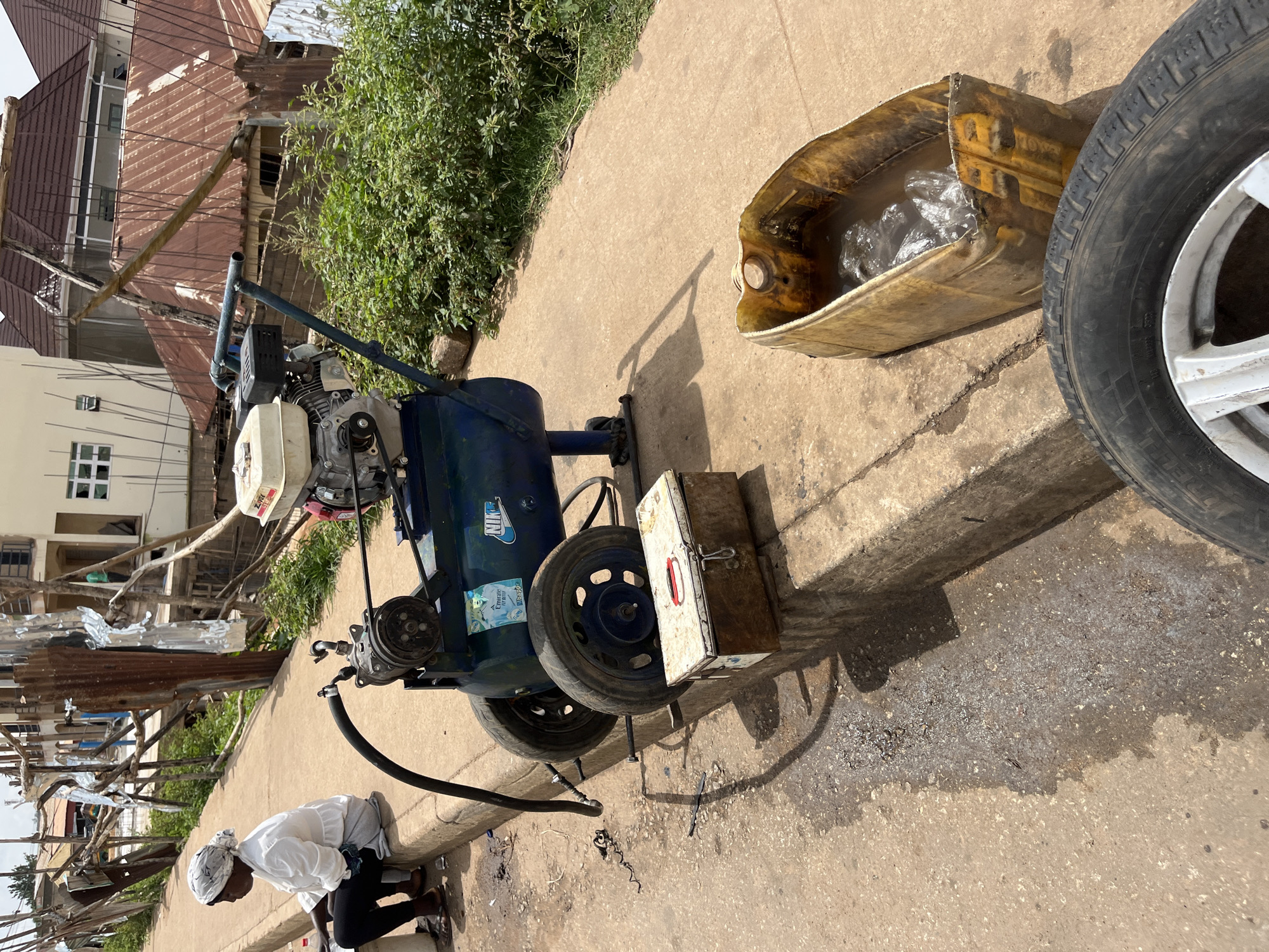 In Bukuru, Nigeria, a scene unfolds on a dusty, sun-drenched roadside. A woman, dressed in a white top and dark trousers, sits on a concrete curb, her head covered with a white scarf. Beside her, a portable air compressor, a piece of machinery with a blue tank and a gasoline engine, rests on two wheels. A black hose snakes from the compressor, its end resting on the ground. Nearby, a rusty metal toolbox sits on the curb, and a yellow plastic container, half-filled with murky water and some plastic bags, lies on the ground. A detached car tire and rim are also visible in the foreground. The background depicts a construction site with unfinished buildings, corrugated iron roofs, and scaffolding, all under a bright, cloudy sky. The environment suggests an ongoing infrastructure development or repair project in a bustling urban setting.
