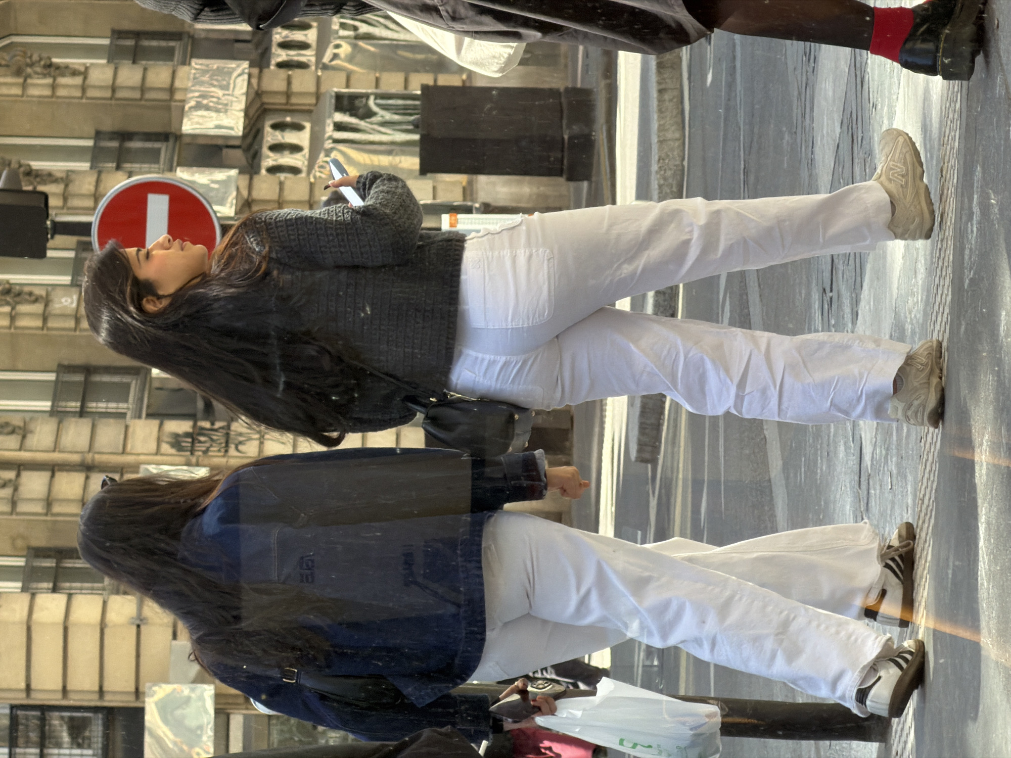 A medium shot depicts two women walking down a sidewalk in Paris, France, as seen through a shop window. The woman nearest the viewer is wearing a dark blue denim jacket over white pants and white sneakers with black stripes. The woman farther away wears a grey sweater over white pants and light tan sneakers. Each woman is holding a phone. The background shows the facade of a typical Parisian building. A red and white no-entry sign is mounted on a pole.