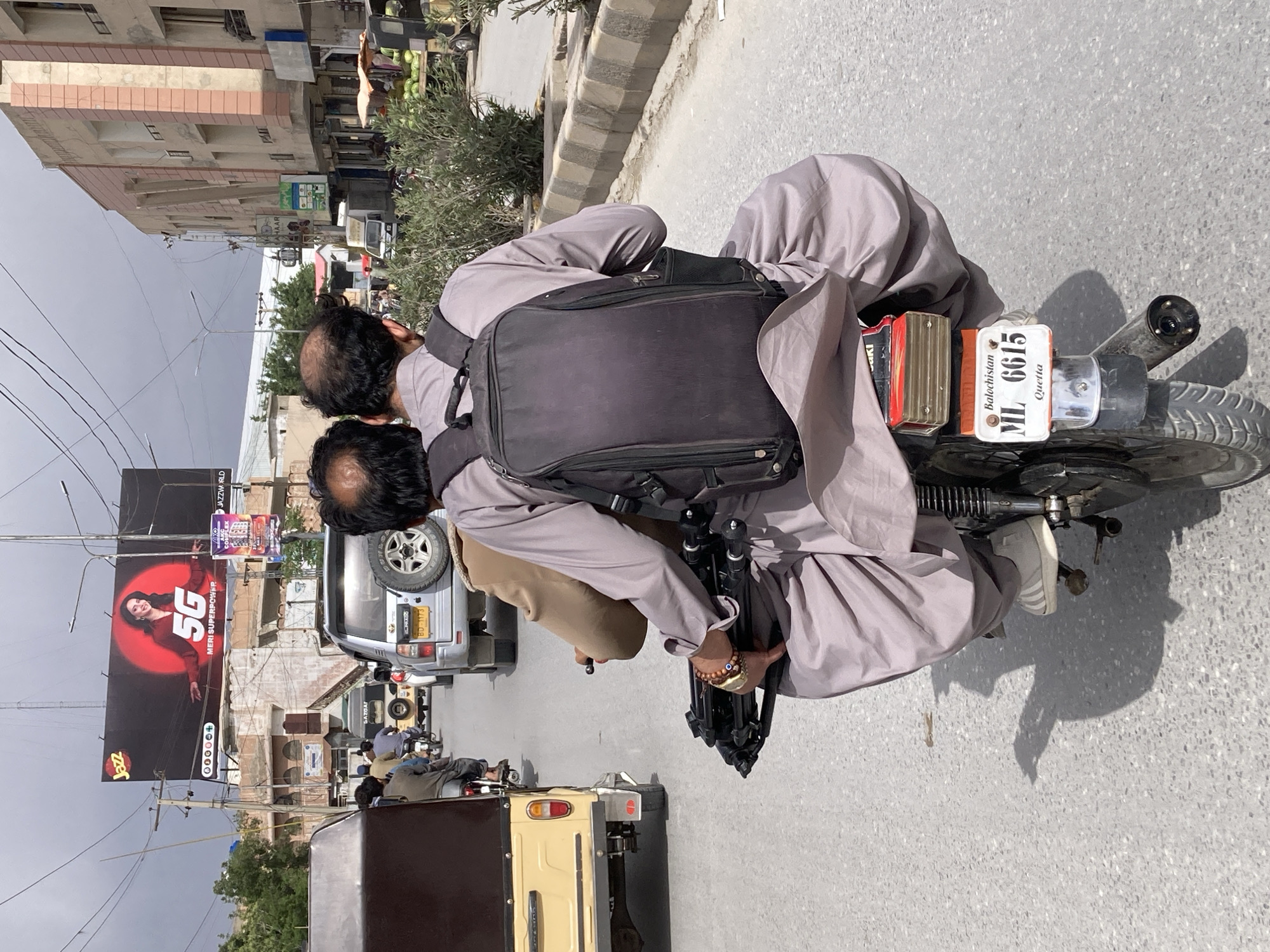 Two men are captured riding a motorcycle on a paved street in Quetta City Tehsil, Pakistan, under an overcast sky during the daytime. The image is oriented vertically, but the subjects are moving horizontally across the frame. Both men appear to be wearing traditional light-colored shalwar kameez attire. The man in front has a black backpack on his shoulders, while the man in the back is holding a black camera tripod.
The motorcycle's rear license plate is clearly visible, reading "Balochistan ML 6615 Quetta," which confirms the location. Other vehicles are present on the street, including a silver SUV with a spare tire mounted on the back, displaying a license plate "BD-1173." Another vehicle in the background has "GAZGAT" written on it.
In the background, there are multi-story buildings and utility poles with numerous power lines crisscrossing the sky. A large billboard prominently features a woman and the text "Jazz," "5G MERI SUPERPOWER," and smaller text "JAZZWORLD" along with "400000." Another smaller sign shows the text "HAAR." The street appears to be busy, reflecting typical urban life in the area.