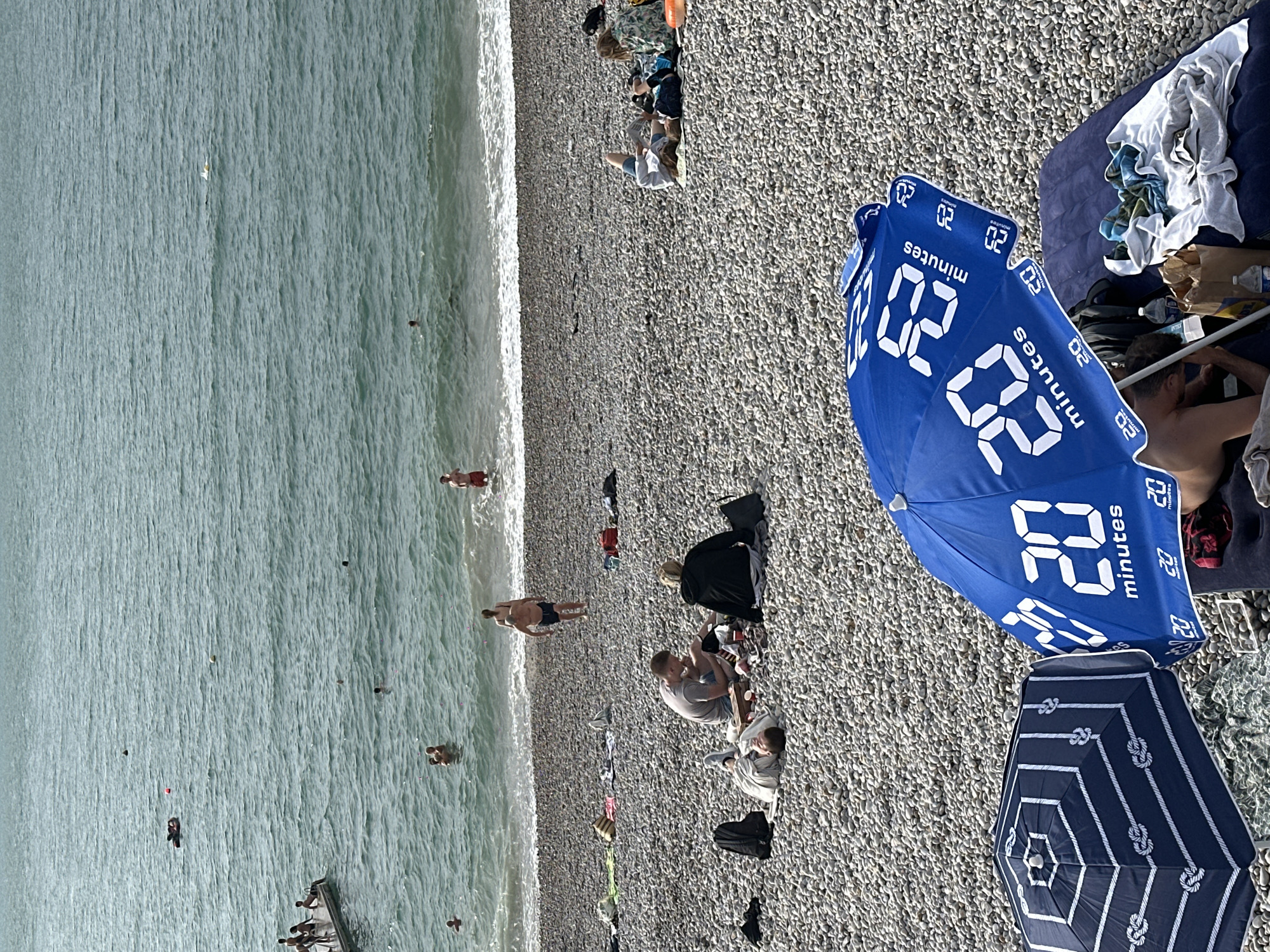 The image depicts a rocky beach in Étretat, France, with people enjoying a sunny day. In the foreground, two umbrellas are set up. One is blue with white lettering that reads "20 minutes" in a digital font, and it is open, providing shade for a person lying on an air mattress underneath. The other umbrella is dark blue and white with a knot pattern.
In the middle ground, several groups of people are visible on the beach and in the water. Some are picnicking on blankets, while others are wading or swimming in the sea. Further back, more people are enjoying the water, with a dock visible on the left side of the image where several individuals are standing. The sea itself is calm with gentle waves. The overall scene conveys a relaxed atmosphere of a summer day at the beach.
