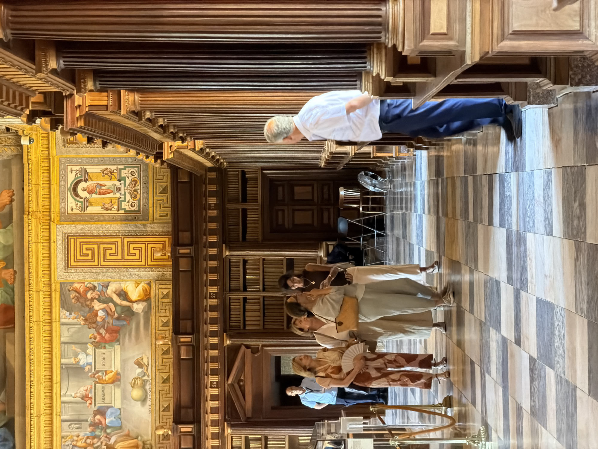 The image depicts the interior of the Royal Library of San Lorenzo de El Escorial, Spain. The vast hall features towering wooden bookshelves lining the walls, adorned with classical paintings and intricate golden architectural details. A man in a white shirt and blue pants stands facing away from the camera, observing the library's grandeur. In the foreground, a group of four women are engaged in conversation and observation. One woman holds a fan, another carries a tan handbag, and a third, wearing glasses, consults her phone. The floor is paved with a striking checkered pattern of light and dark grey marble tiles. The overall scene conveys an atmosphere of scholarly retreat and historical significance.