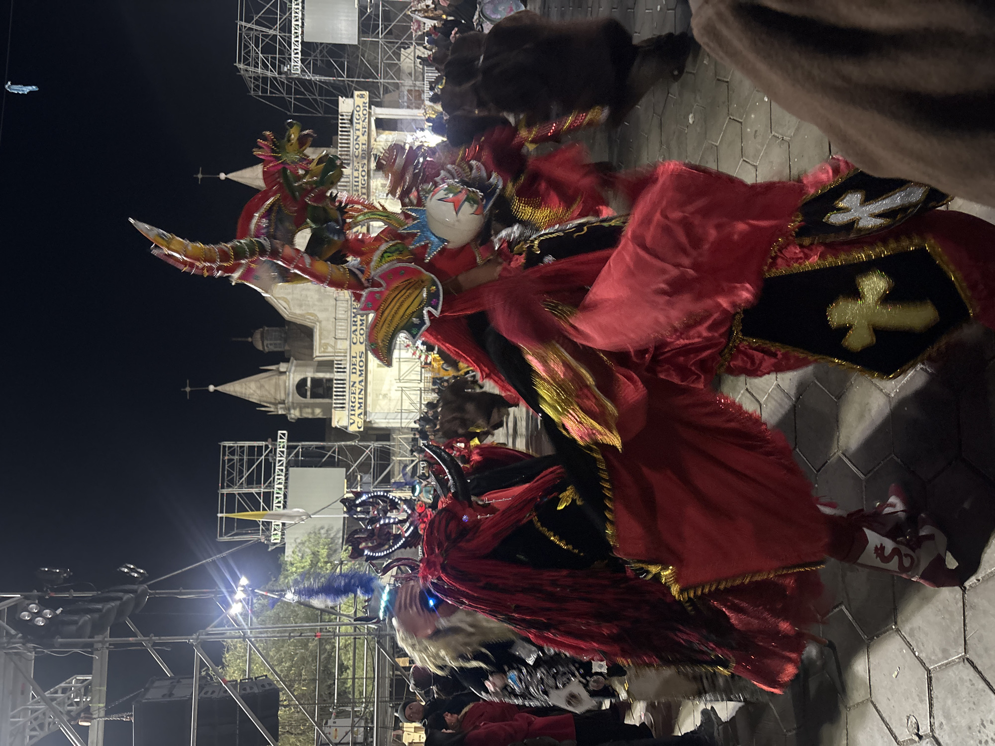 The image captures a nighttime street scene in Pozo Almonte, Chile, featuring a procession of individuals in elaborate costumes. The foreground is dominated by two figures in ornate, vibrant attire, one in predominantly red with flowing fabric and gold trim, and another with a striking headdress adorned with blue feathers and horns. The costumes appear to be inspired by traditional folkloric or religious celebrations, with one of the figures wearing boots decorated with a serpentine motif. In the background, a church with a prominent cross on its steeple is visible, illuminated by artificial lights. A banner displaying text, partially obscured but including the phrase "VIRGEN DEL CARMEN", suggests a religious context. The scene is set against a dark night sky, with scaffolding and lighting rigs indicating a staged event or festival. A crowd of onlookers can be seen further in the background, observing the procession. The ground is paved with hexagonal stones.