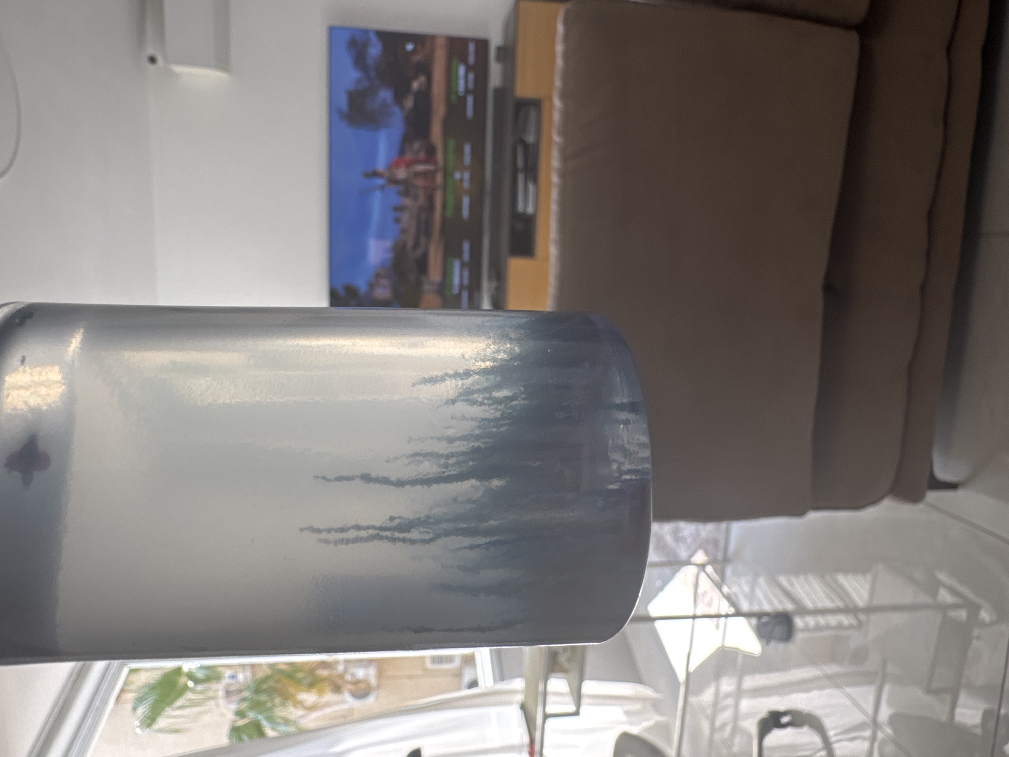 An indoor shot shows a cylindrical object with a textured, possibly metallic surface, in the foreground, obscuring the view of the background. The object has a gradient of colors, from light blue at the top to darker blue at the bottom, with vertical, branch-like patterns. In the background, a large television screen displays a video game. To the left of the television, a wooden cabinet is visible. A light-colored couch occupies the right side of the frame. The environment appears to be a living room, with bright light coming from a window on the left side of the image, suggesting a daytime scene. The style of the television stand and the overall aesthetic suggest a modern home. The overall impression is that of a candid shot, possibly taken at a residence in Netanya, Israel.