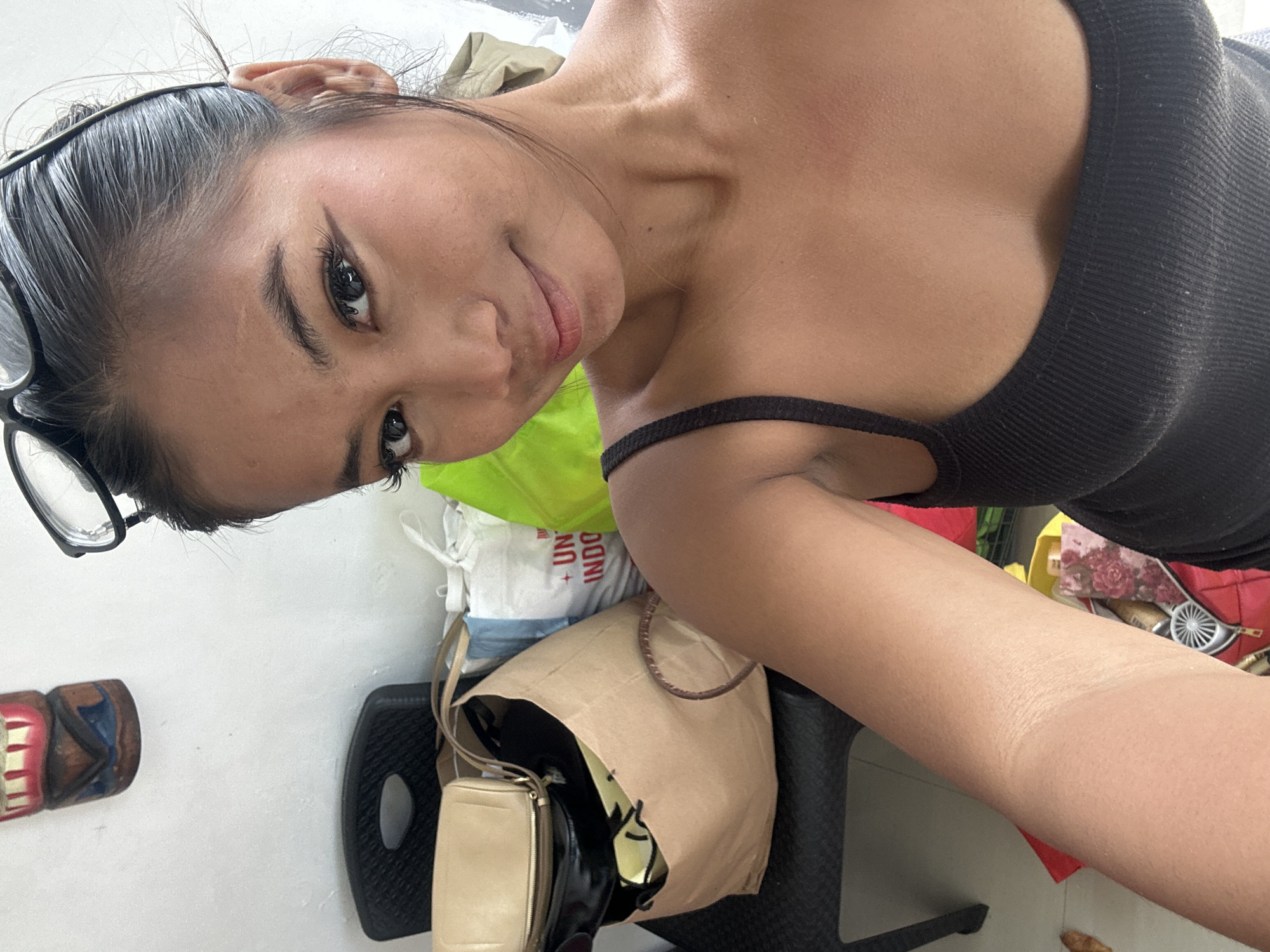 A young woman with dark hair, pulled back and wearing glasses on top of her head, poses for a selfie in what appears to be a room in Sukawati, Indonesia. She is wearing a black tank top. To her left, a black plastic chair supports several bags, including a large brown paper bag and a cream-colored clutch. Behind the bags, a white bag with red lettering that reads "UN INDO" is visible. A portion of a green bag is also present. A decorative wooden carving, possibly a totem or mask, hangs on the white wall in the upper left corner of the image. The lighting suggests an indoor setting, possibly a home or guesthouse.