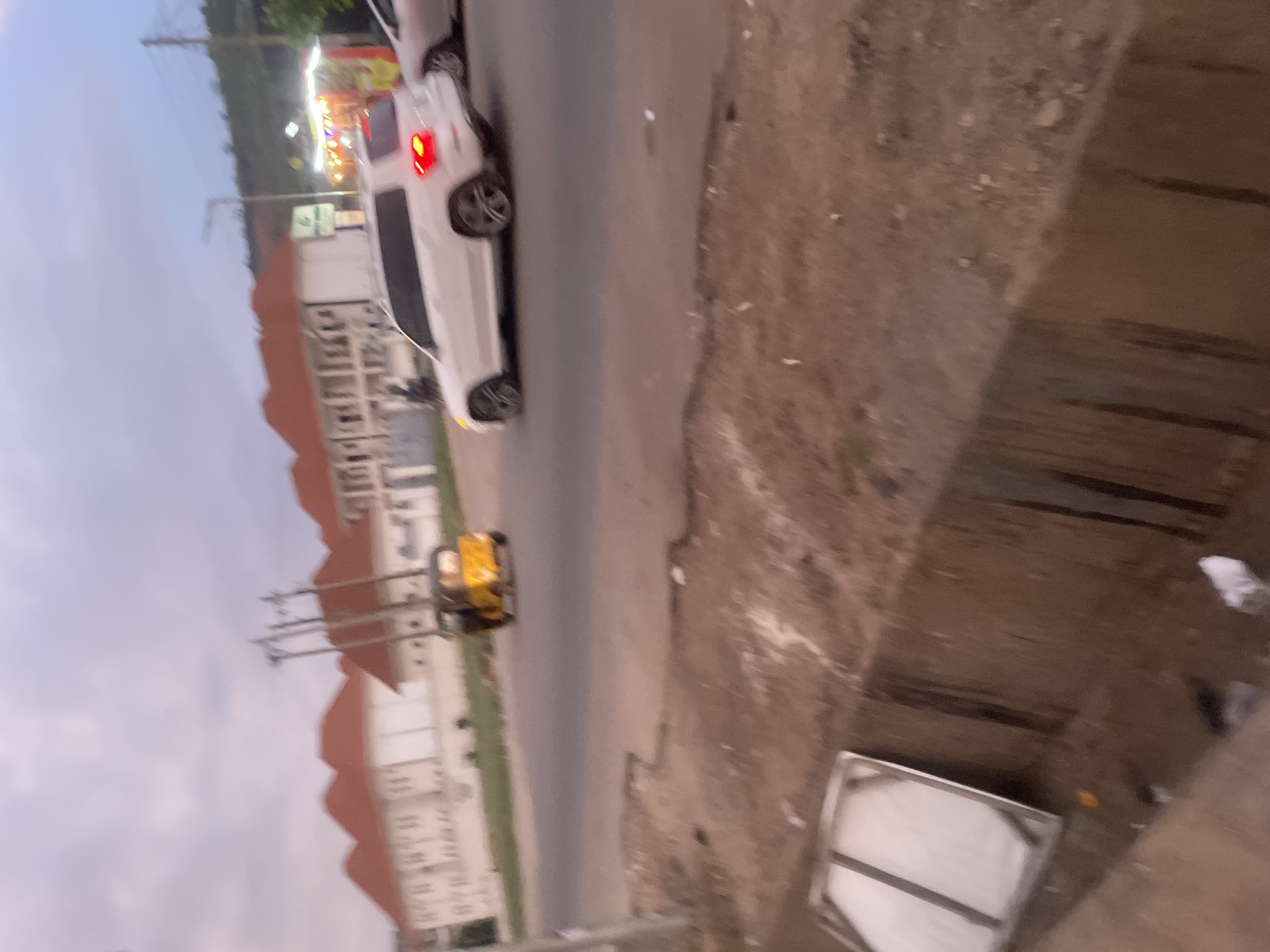The image shows an urban street scene in Abuja, Nigeria. A white car is driving on the road. There's a yellow generator near the road. Buildings are visible in the background. The pavement and the road are visible. A metallic square object is seen near the edge of the pavement. There is vegetation or foliage visible in the distance.