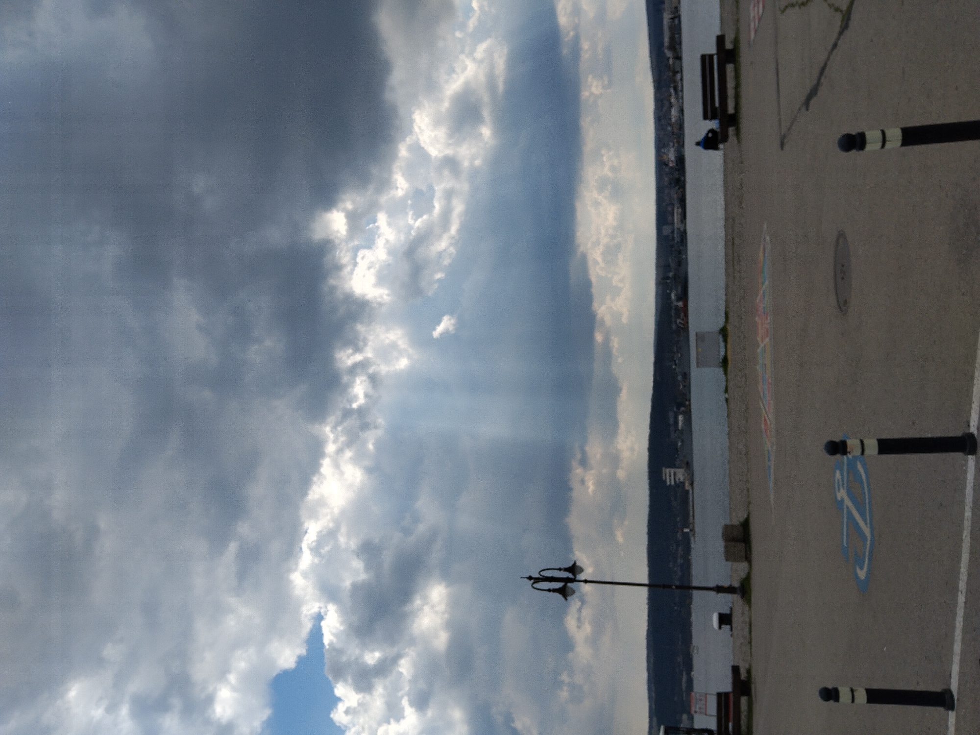 The image shows a waterfront scene in Varna, Bulgaria. The sky is filled with dramatic clouds, some allowing sunlight to stream through. The waterfront promenade includes a concrete area with black and white striped bollards, a lamppost, benches, and painted parking spaces, including one with a handicapped symbol. The water is visible along the edge, and a tree-covered shoreline rises in the distance. A single person is seated on a bench.