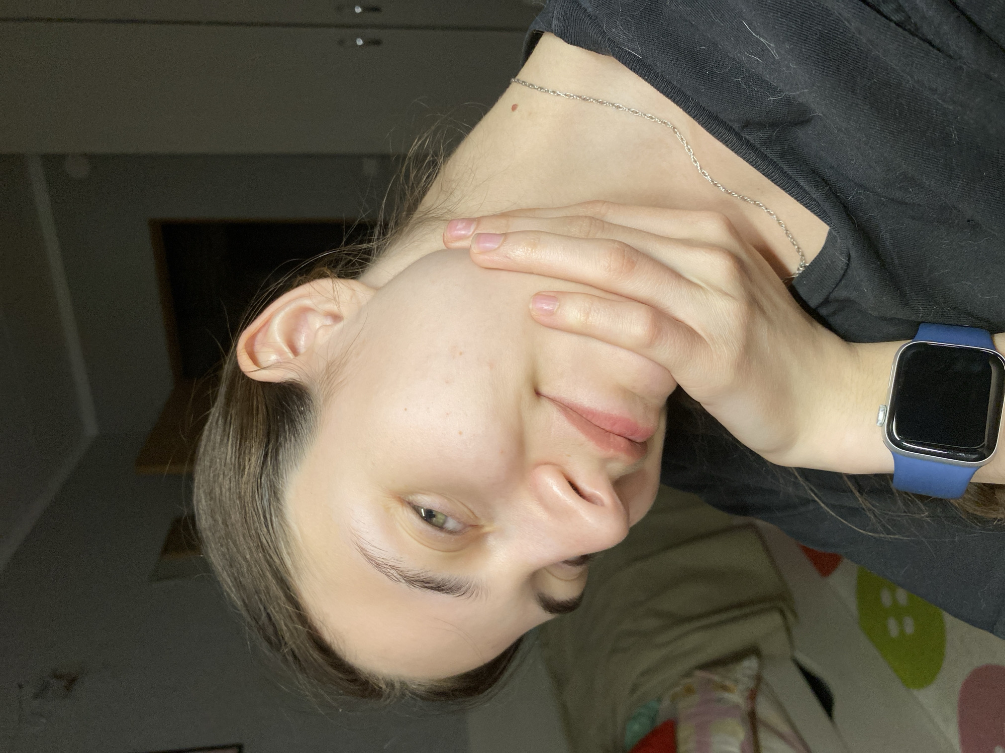 A close-up image shows a woman with fair skin and brown hair, wearing a black t-shirt and a silver chain necklace. She is wearing a smartwatch with a blue band on her left wrist. Her left hand is resting on her cheek. The background is blurry and indistinct, but includes a glimpse of a wooden structure and a surface with green and pink shapes. No information in the image indicates the location is Votkinsk, Russia, or any specific city.