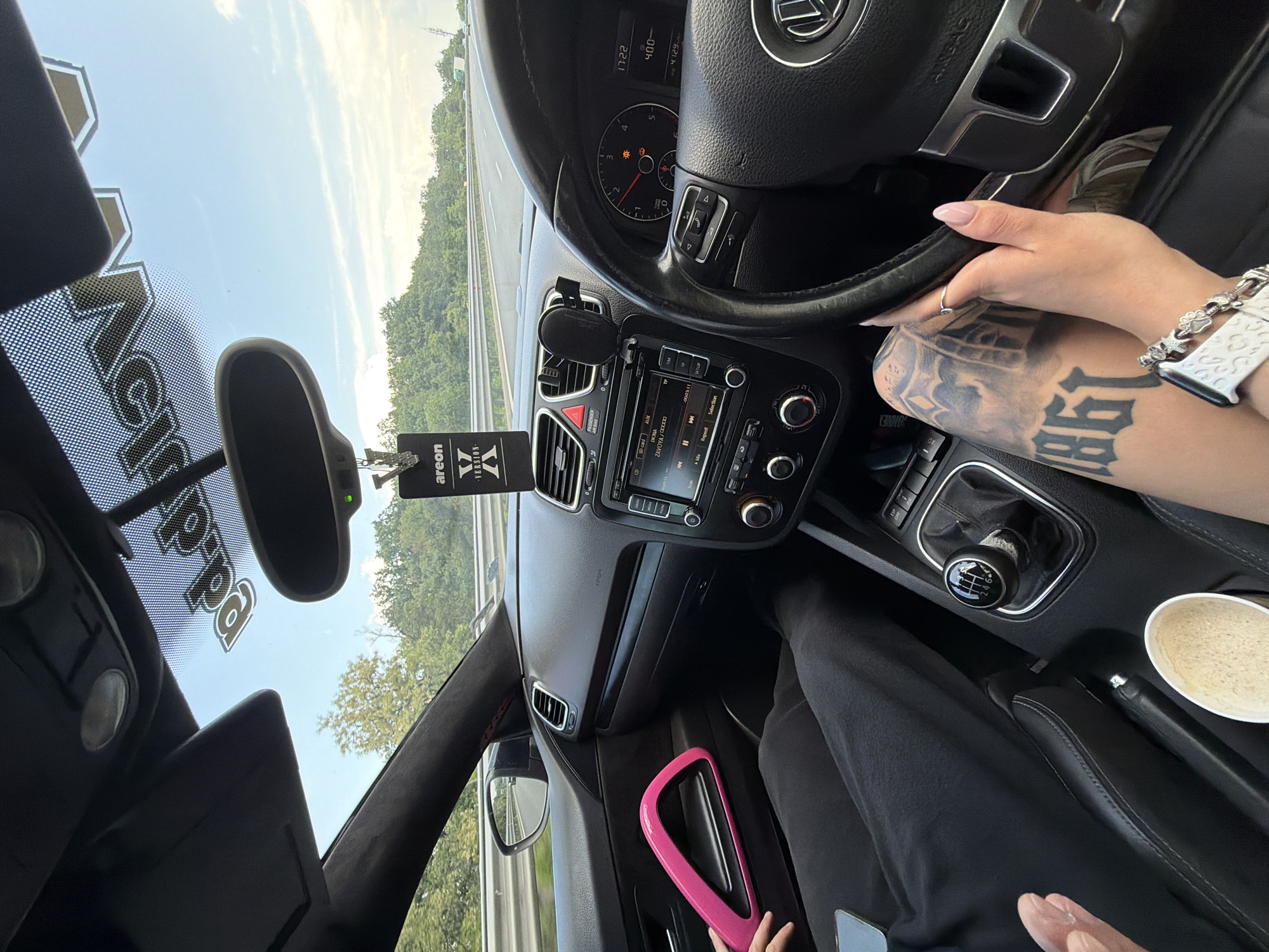 The image depicts the interior of a Volkswagen vehicle, consistent with a Scirocco (Mk3), in motion on a multi-lane asphalt highway near Novi Han, Bulgaria, under a clear blue sky.
The perspective is from the front passenger seat, looking forward and towards the driver's side. The driver's left hand, adorned with light pink polished nails and a white smartwatch with a silver charm bracelet (featuring paw print charms), rests on the leather-wrapped steering wheel, which bears the Volkswagen logo. Visible tattoos, including the numbers "1961" and other designs, are present on the driver's left leg/thigh area. The instrument cluster shows the time as "17:22" and a temperature reading of "4.0" or "40.0", with an illuminated warning light. The vehicle is a left-hand drive model with a 6-speed manual transmission, indicated by the gear knob.
The front passenger's legs, clad in black trousers, are visible. The passenger's right hand holds a smartphone, partially obscured, resting on their lap. A white paper cup containing a frothy beverage is positioned in the center console. The front passenger door panel features a distinct bright pink grab handle/trim.
The dashboard includes a factory infotainment system displaying "ZAVOYA /////" and a CD card/AUX input. A black phone mount is affixed to the dashboard. An "Areon Version X" air freshener hangs from the rearview mirror. A "Vеlopa" sticker is visible, applied upside down, on the upper part of the windshield.
Outside the vehicle, the highway is bordered by a metal guardrail and densely wooded areas with green deciduous trees. Other vehicles are visible in the distance on the multi-lane road.
