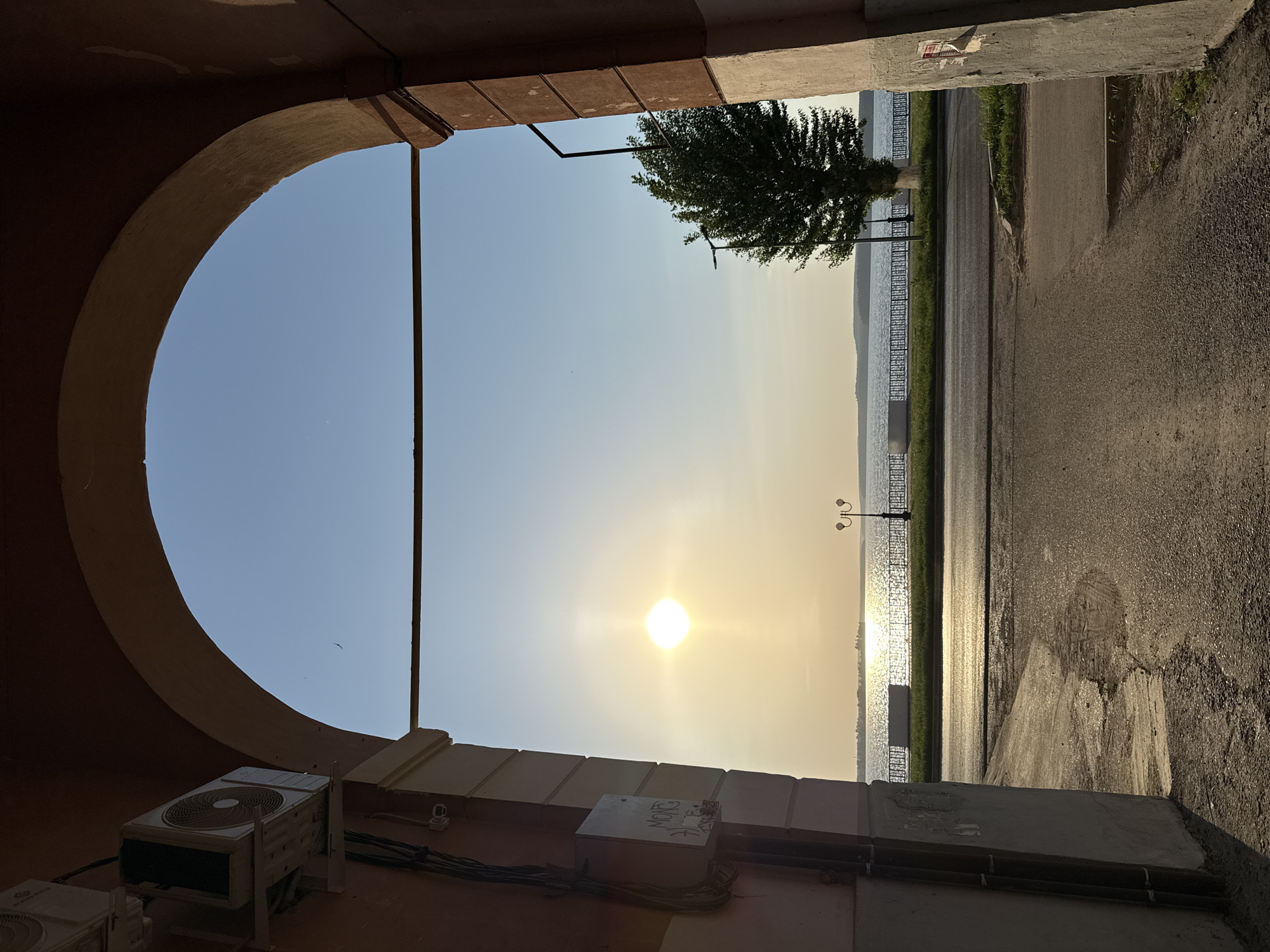 A partially obscured view of a riverside landscape is framed by an arched, building entryway. The sun is low on the horizon, reflecting strongly on the water’s surface. A tree stands on the shoreline, next to a decorative fence. A paved area extends towards the viewer. Parts of the building’s exterior, including air conditioning units and electrical boxes, are visible in the foreground, indicating an urban setting. The scene likely depicts the waterfront in Votkinsk, Russia.