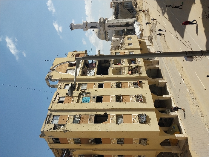 The image depicts a scene of extensive urban damage in Khan Yunis, Palestinian Territory, under a clear blue sky with scattered white clouds. A prominent multi-story residential building, light brown with darker brown banding and white decorative window accents, occupies the left and central midground. This building exhibits widespread damage, including numerous impact holes, shattered windows, and significant structural compromise to its upper floors and roof. Debris, including clothing and blue fabric, hangs from multiple windows and balconies. The ground floor features a series of arched openings, beneath one of which a light green vehicle is partially visible.
A paved street runs in front of the damaged building. On this street and an adjacent paved walkway, approximately ten individuals are visible. One person in dark clothing walks on the pavement in the lower-left. Further down the street, another individual in a red shirt walks, while several smaller figures, consistent with children, are present further in the distance, some appearing to play with a ball on the roadway.
To the right of the residential building, beyond the street, lies a large expanse of rubble and demolished structures, within which at least one white tent-like shelter is erected. In the background, a mosque featuring a white dome and a tall, slender white minaret stands partially damaged, with visible structural compromise to the minaret's upper section. Additional heavily damaged or collapsed buildings are discernible beyond the mosque. A grey utility pole with multiple wires extends vertically on the right side of the frame, with a street light fixture attached to a similar pole in the mid-left.