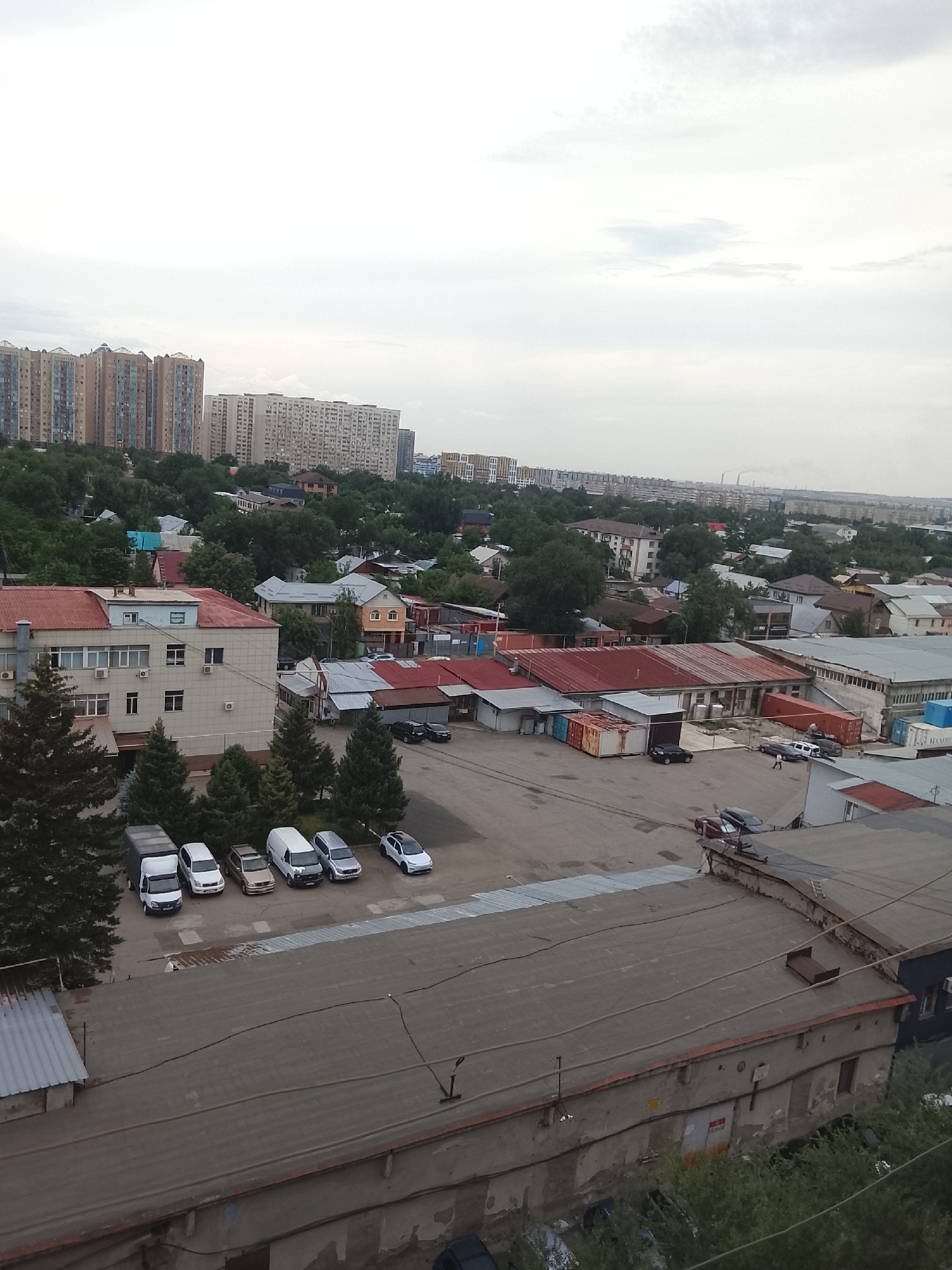 The image shows a panoramic view of an urban area with high-rise buildings, residential houses, and industrial structures. In the foreground, there is a rooftop with a concrete surface, and part of a low-rise building with peeling paint. The midground features a courtyard with several parked vehicles, including vans and cars, and a building with a red-tiled roof. Further back, there are more residential buildings and a cluster of tall apartment blocks. The background reveals a hazy skyline with more buildings and what appear to be industrial chimneys. The sky is overcast, suggesting a cloudy day. The presence of various architectural styles indicates a mixed-use urban environment. The scene appears to be a residential and industrial district within the city of Almaty, Kazakhstan, possibly in the eastern part of the city, given the landscape and building styles common in Almaty.