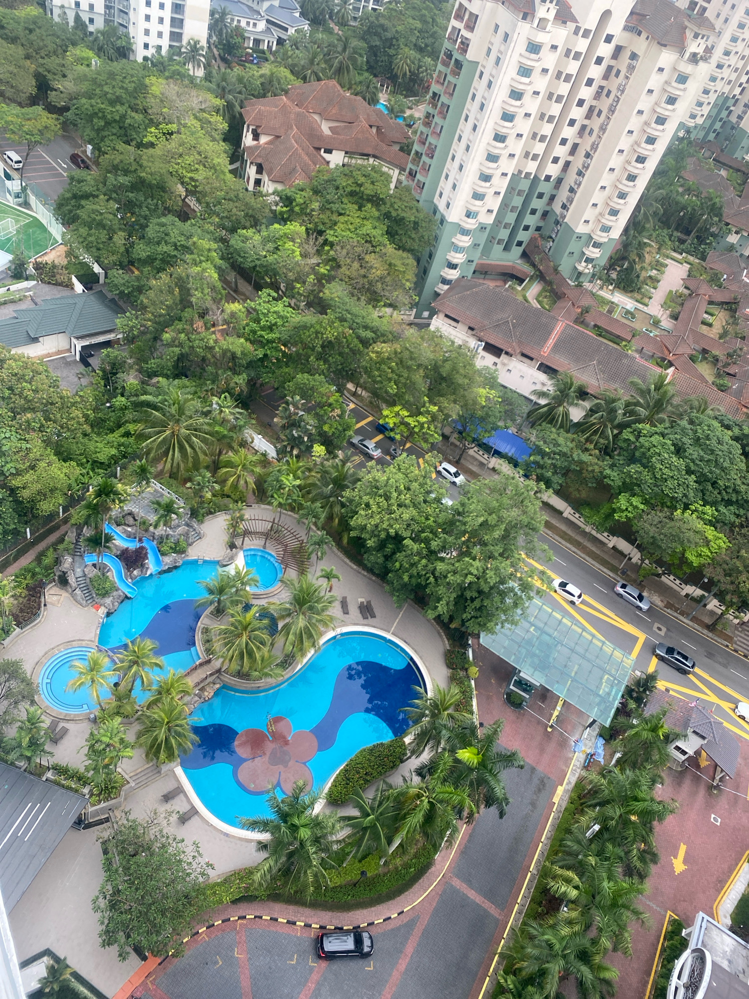 This is an aerial view of a residential complex in Kuala Lumpur, Malaysia, likely in a tropical or subtropical climate given the abundant greenery. The focal point is a large, uniquely shaped swimming pool with bright blue water, featuring a smaller circular pool connected by a bridge. One section of the main pool has a large, stylized red flower design. Adjacent to the pool is a water slide complex with multiple blue slides and rocky landscaping. Surrounding the pool area are palm trees and other lush vegetation.
In the background, several multi-story apartment buildings with light-colored facades and numerous balconies are visible, interspersed with smaller, terra-cotta-roofed buildings that suggest a lower-density housing area or older structures. A road runs alongside the complex, with several cars visible, including a black SUV on a paved circular driveway at the bottom right and other vehicles on the road. There is a covered entrance or parking area with a glass roof. The overall impression is one of a well-maintained, amenity-rich residential environment within a densely vegetated urban setting.