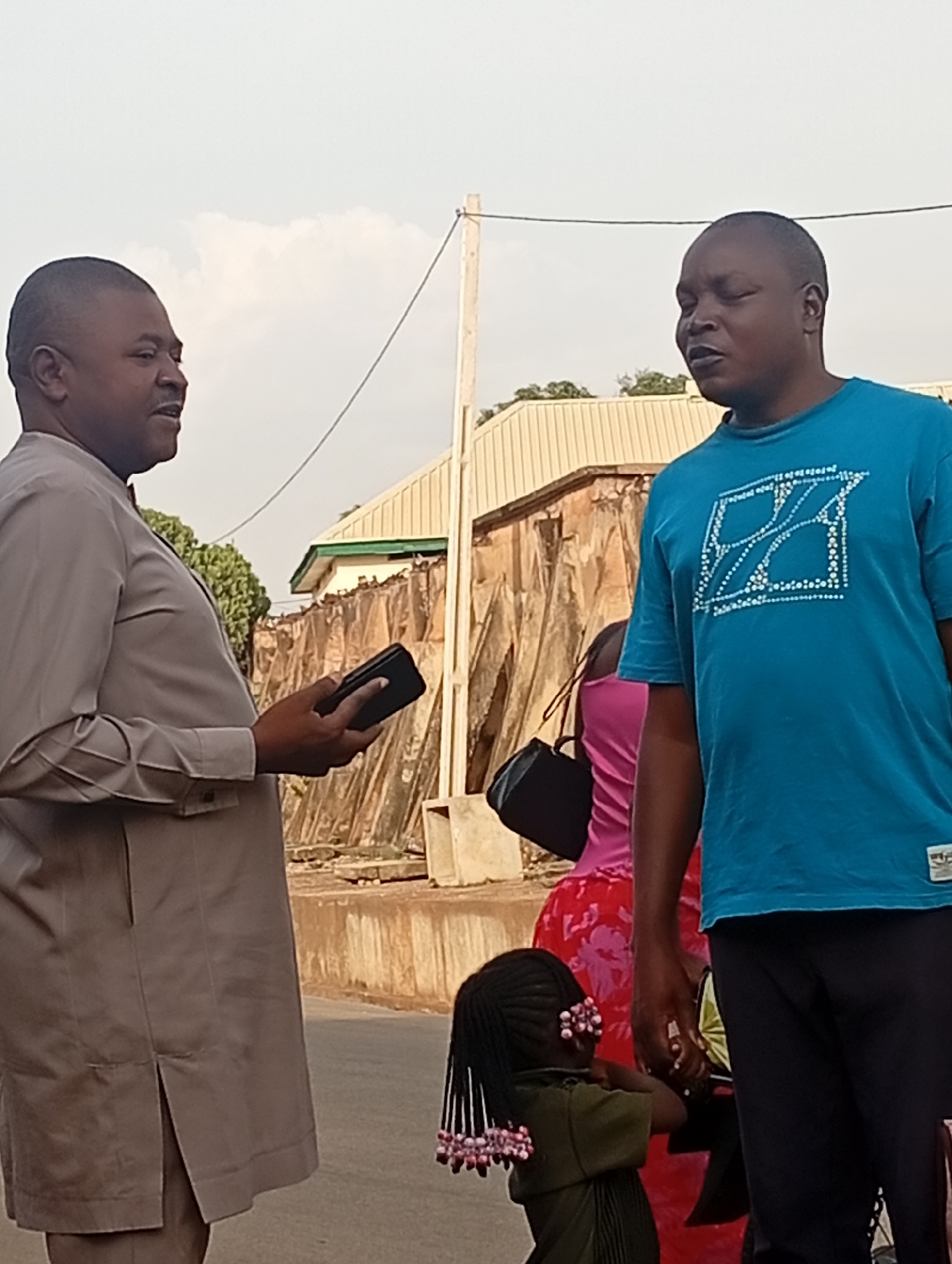 Two men are standing on a street in Gura Top, Nigeria, engaged in conversation. The man on the left is wearing a gray tunic and holding a black object, while the man on the right is wearing a blue T-shirt and black pants. Behind them, there are other people, including a child with decorated braids. The street has buildings and utility poles in the background.
