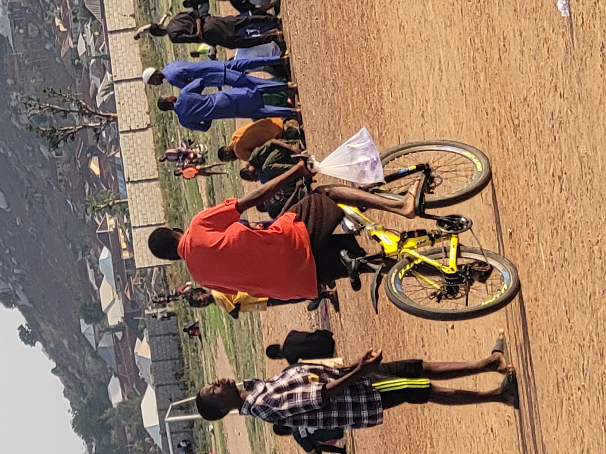 The image depicts a group of people, potentially school children, gathered in an open dirt field on the outskirts of the city of Ibo, Nigeria. A boy in a red shirt is sitting on a yellow bicycle holding a plastic bag. Other individuals are standing in a row. The background shows residential buildings and a hill. A goal post suggests the field is used for sports. Some of the children are wearing blue uniforms.