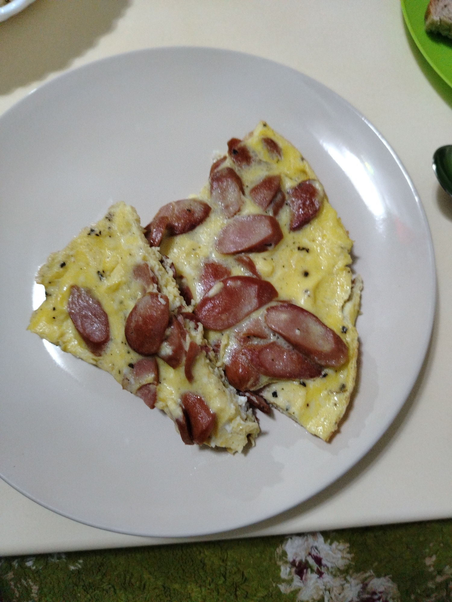 The image shows two slices of an omelet with sausage on a white plate. The omelet is yellow with visible black specks and slightly browned edges. Sliced sausage pieces are arranged on top. The plate sits on a table with a patterned tablecloth. The location is unknown and there are no people visible in the image.