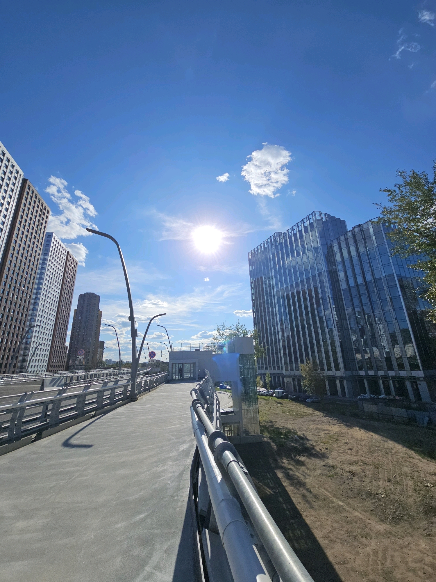 This is a wide-angle, eye-level shot taken on a bright, sunny day, likely in the city of Moscow, Russia. The perspective is from a pedestrian walkway or bridge, looking forward and slightly to the right. The walkway is made of light gray concrete with a metal railing on both sides. Streetlights with curved poles are visible along the walkway.
In the background, several tall, modern buildings dominate the cityscape. To the left, there are two high-rise buildings with a grid-like facade pattern, one appearing to be made of brown bricks and the other of lighter colored material. To the right, a striking glass-fronted building stands out, its reflective surfaces mirroring the blue sky and white clouds. A lone figure is visible in the distance, walking into an entrance of a building on the right side of the walkway.
The sky is a vibrant blue, with scattered white cumulus clouds. The sun is bright and high in the sky, casting long shadows from the walkway's railing and streetlights onto the concrete surface. The area beside the walkway features a grassy, somewhat unkempt embankment. The overall impression is one of modern urban development under a clear sky.