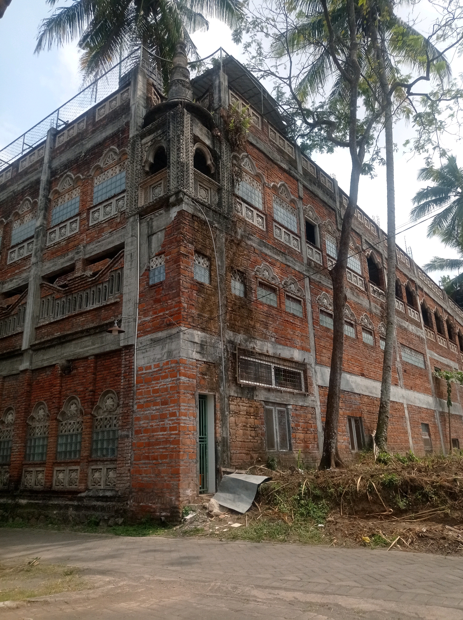 The image shows a tall, multi-story brick building with ornate architectural details. The building appears aged, with some signs of wear and tear on the brickwork and concrete elements. The windows are a mix of glass blocks and framed panes, many with decorative surrounds. Several trees, including palm trees, are growing around the building, with some branches reaching over the structure. A metal fence runs along the top of the building. The ground in front of the building is a paved surface, possibly a road or walkway, with some dry vegetation and debris. The sky is visible in the upper portion of the image, appearing overcast or hazy. The scene is situated in an outdoor environment, likely a non-urban or semi-urban area given the presence of mature trees and less formal landscaping. The image is a static shot, with no people or animals visible. The overall impression is one of an old, possibly abandoned or neglected, building in a tropical or subtropical setting, consistent with locations like Sananrejo, Indonesia.