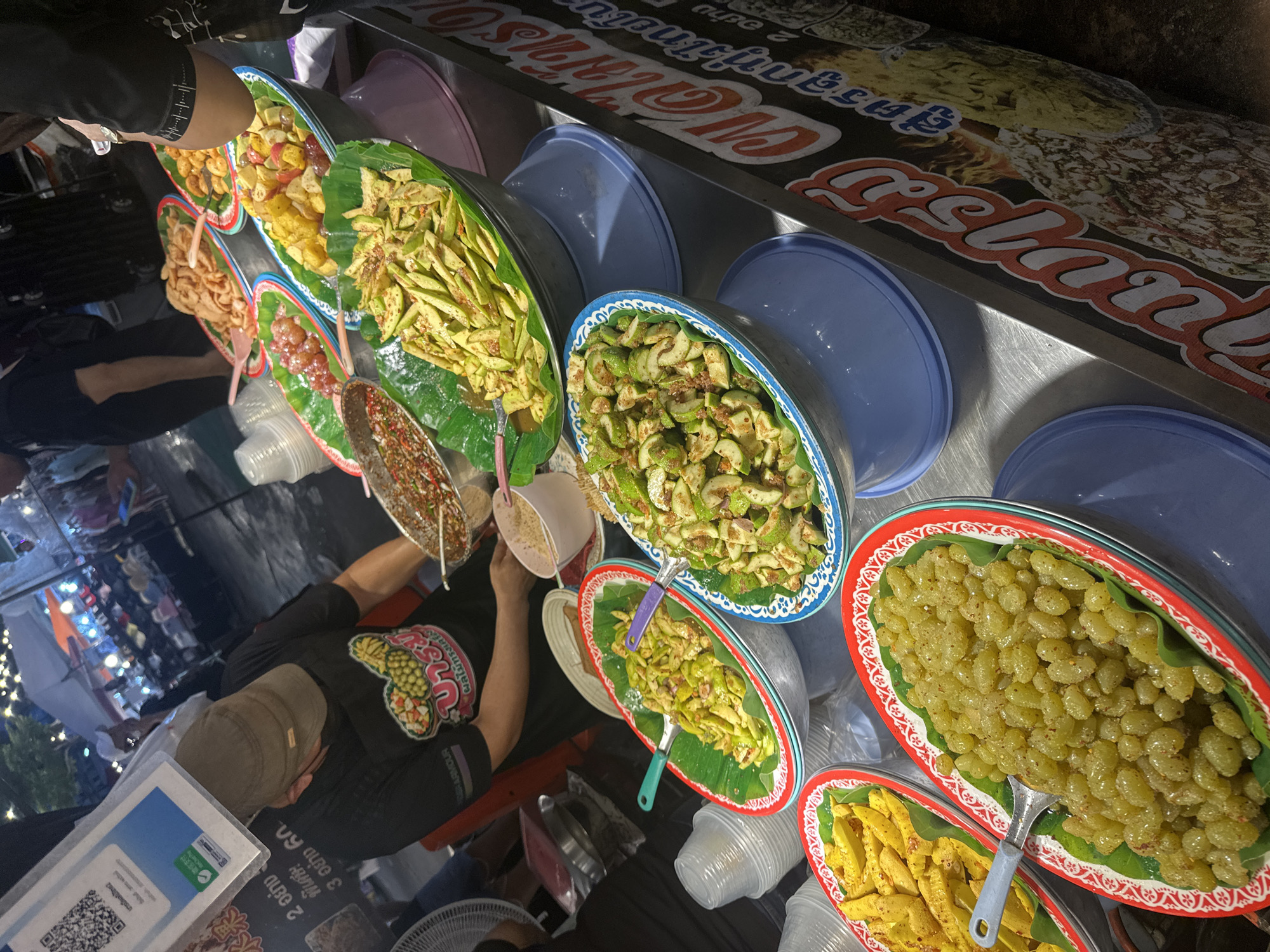 The image depicts a bustling street food stall in Phuket City Municipality, Thailand, likely at night given the artificial lighting. A vendor, wearing a black t-shirt and a brown cap, is seen preparing food. The stall displays a variety of colorful fruits and snacks, served in large metal bowls lined with banana leaves, and presented on a metal counter. In the foreground, a bowl filled with small, green, grape-like fruits is prominent, with a spoon resting in it. Adjacent to it, a bowl of sliced green fruits, likely unripe mangoes, is also visible. Further back, other bowls contain an assortment of prepared fruits and snacks, some appearing fried or pickled. The background suggests an outdoor market setting with tents, lights, and other stalls. A QR code and some Thai signage are also visible. The overall scene captures the vibrant atmosphere of a Thai street food market.