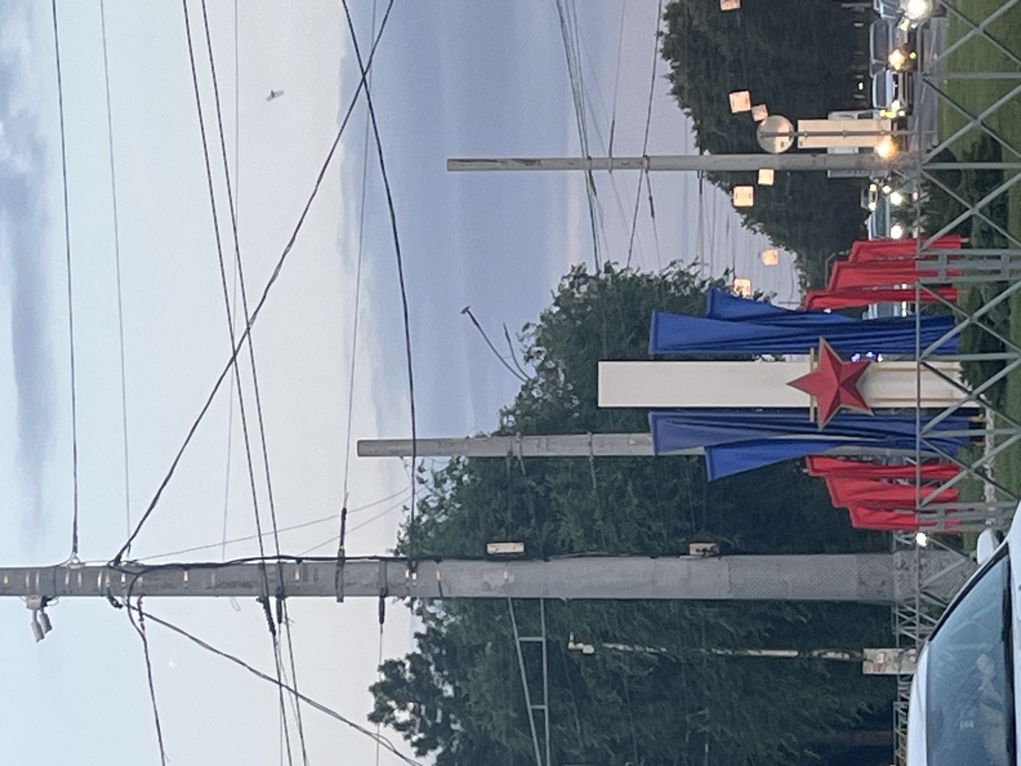 The image captures a street scene in Krasnodar, Russia, likely during the evening or overcast conditions. In the foreground, the hood of a white car is visible on the left. A metal fence runs horizontally across the lower part of the image. Behind the fence, a patriotic-themed display features a red star on a white pedestal, flanked by draping red and blue fabrics. In the mid-ground, trees form a dense green backdrop. Various utility poles with numerous wires crisscross the frame, one on the far left with two cameras mounted near its top. A second, thinner utility pole stands to the right of the display. Beyond these, a street with traffic is evident, with cars and streetlights illuminated. Several square decorative lights are suspended from wires overhead in the distance. The sky is cloudy and a pale blue.