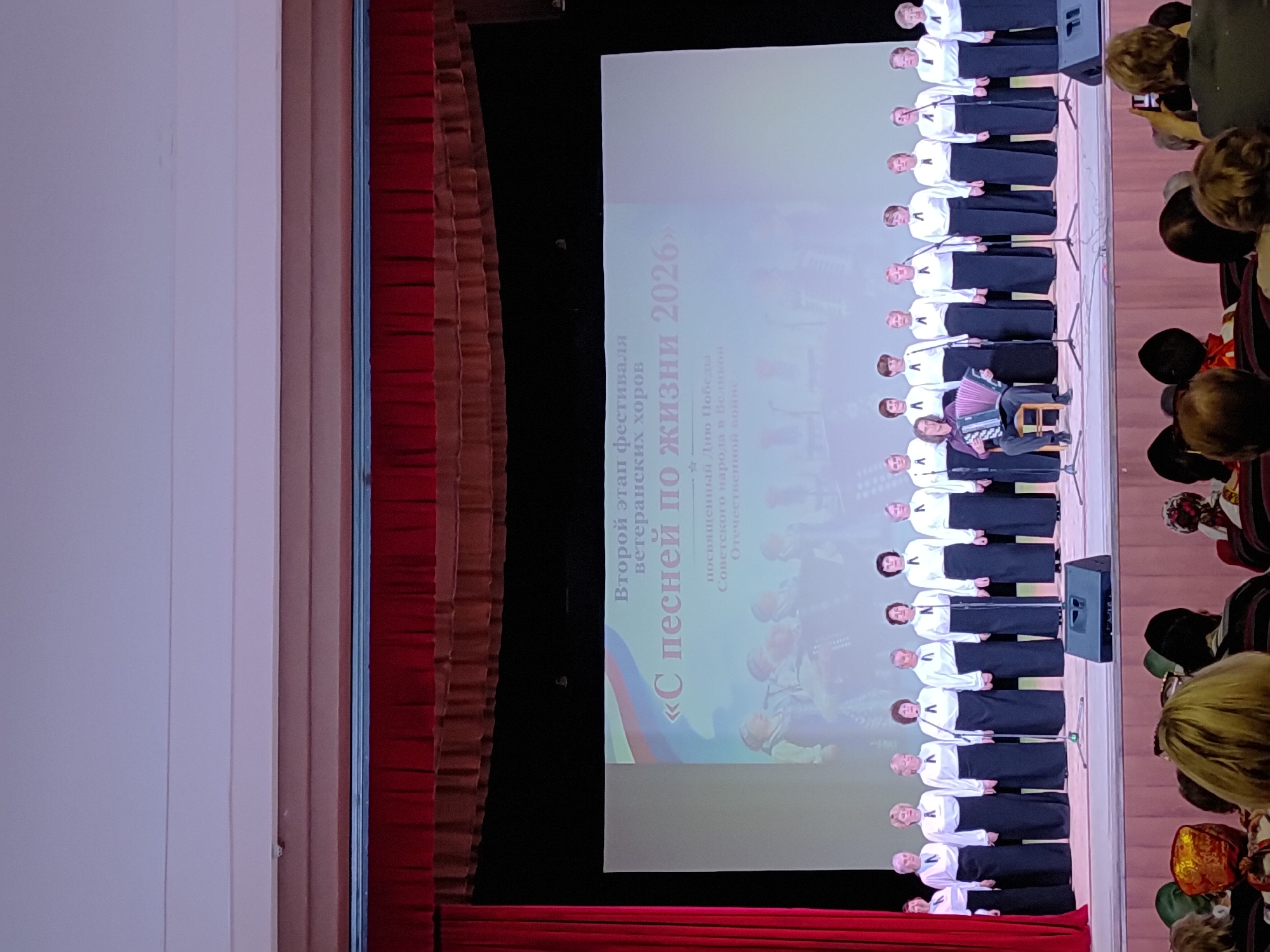 Here is a detailed description of the image:
**Scene:** The image captures a performance on a stage, likely in a theater or auditorium.
**Subjects:**
* A choir of elderly women are standing in rows on the stage. They are dressed in similar attire - white tops and dark skirts.
* An individual sits on a stool with an accordion.
* Audience members are visible in the foreground.
**Objects:**
* Microphones stand in front of each row of the choir.
* Speakers stand on the sides of the choir.
* A projection screen displays text in Cyrillic script along with images of military scenes in a washed-out manner. The text translates to "Second Stage of the Veteran Choirs Festival" and the title, "With a Song Through Life 2026," dedicated to Victory Day.
* The stage has red curtains along the edges.
**Actions and Interactions:**
* The choir appears to be singing or preparing to sing.
* The accordionist is playing.
* Audience members are watching and some are taking pictures.
**Environment:**
* The lighting on stage is bright, focusing on the performers.
* The auditorium appears to be full or mostly full.
**Location (Based on the event):**
* Unknown, Unknown. Given the Cyrillic script and the celebration of "Victory Day", it is probably related to some event in the Russian Federation.