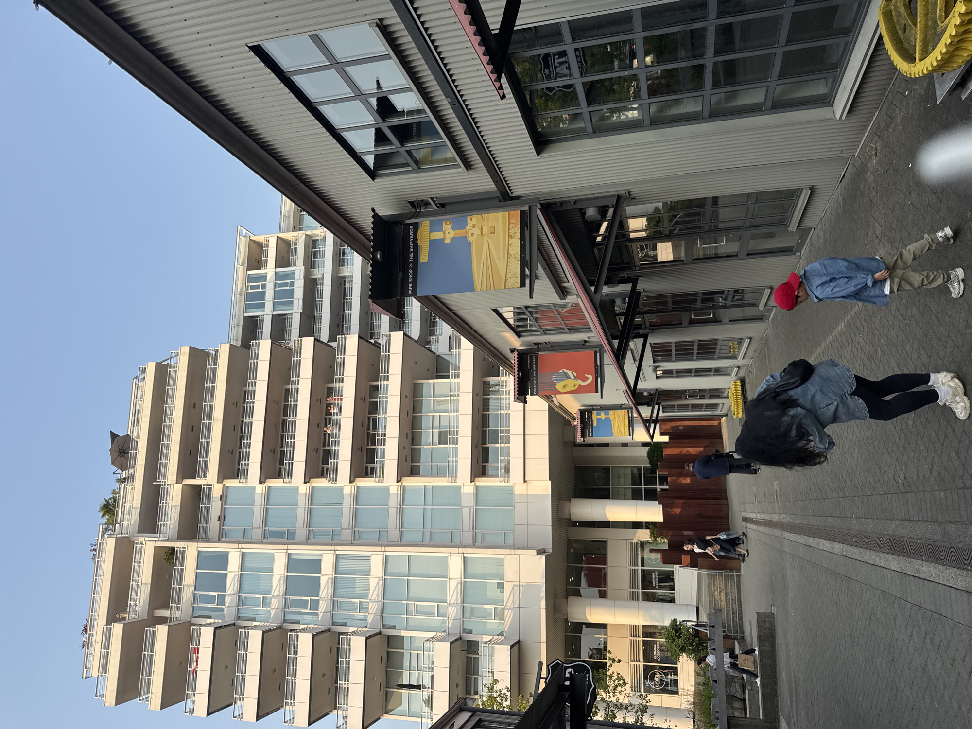 This image depicts a view of The Shipyards district in North Vancouver, Canada, characterized by a modern residential building and a row of industrial-style commercial spaces. In the foreground, a woman with long black hair wearing a denim jacket and black leggings bends over, while a young boy in a red cap and blue jacket walks ahead of her. Further in the background, several other pedestrians are visible, including a man in a dark jacket and a woman in a white shirt. The commercial buildings feature corrugated metal siding and large windows, with signage for "PIPE SHOP @ THE SHPYARDS". The residential building is a tall, multi-story structure with numerous balconies and large glass windows. The ground is paved with interlocking bricks. The scene is bathed in the warm light of a setting sun, casting long shadows.