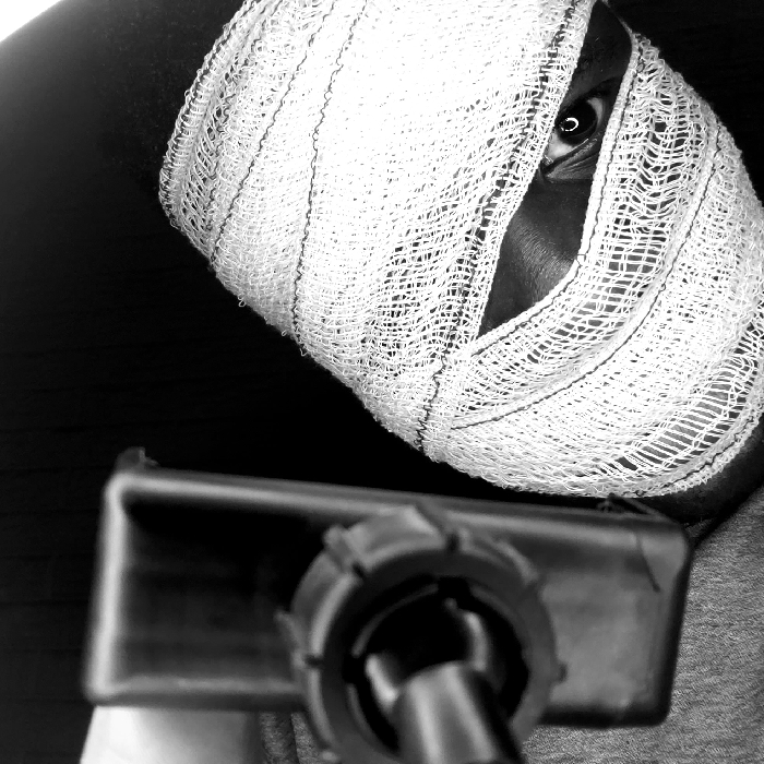 The image is a high-contrast, black and white close-up shot centered on a person's head, which is largely obscured by white medical bandages. The bandages, appearing to be gauze, are wrapped tightly around the head, leaving only a narrow vertical slit through which one dark-colored eye is visible. The eye is open and appears to be looking directly or slightly off-camera, with a distinct catchlight reflecting in its pupil, indicating a source of light. The skin visible around the eye is dark in tone. The texture of the bandages is clearly discernible, showing the interwoven threads and porous nature of the material.
In the foreground, occupying the lower portion of the frame, is a dark, out-of-focus object. This object strongly resembles the barrel and sight mechanism of a firearm, such as a pistol or the front end of a rifle. It is predominantly dark, with some lighter reflections on its metallic or plastic surface, and features a textured, possibly knurled, circular component towards the right, consistent with a gun's barrel or sight adjustment. Its blurred appearance suggests it is very close to the camera lens, creating a shallow depth of field that emphasizes the bandaged head.
The background is uniformly dark, almost black, providing a stark contrast to the white bandages and highlighting the subject. No specific location or time of day is apparent. The overall mood conveyed by the image is one of tension, mystery, or distress, amplified by the monochromatic palette, the concealed face, and the menacing presence of the blurred foreground object. The visible eye peering through the bandages serves as a powerful focal point, suggesting vulnerability, observation, or perhaps a challenging gaze. There is no visible text in the image.