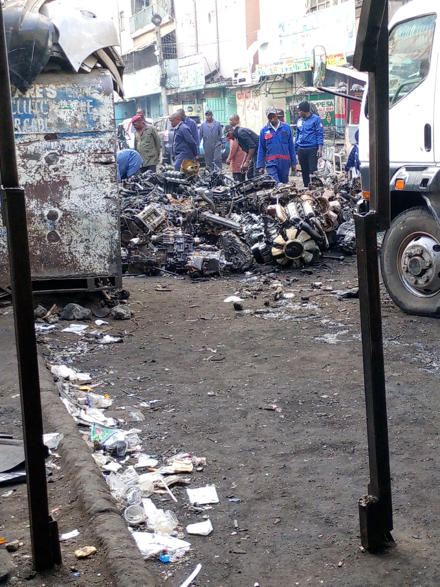 The image depicts a scene in what appears to be a scrapyard or auto parts market in Nairobi, Kenya, given the presence of Swahili signage on nearby shops. Several people are gathered around a large pile of used car engines and mechanical parts. Some individuals are wearing work uniforms or overalls, suggesting they may be mechanics or involved in the salvage and resale of these components. The engines and parts are lying on a dirt or asphalt surface, amidst scattered debris. A truck is parked nearby, possibly used for transporting the parts. The foreground shows trash and litter along the side of the road. The scene suggests an informal and somewhat chaotic urban environment.