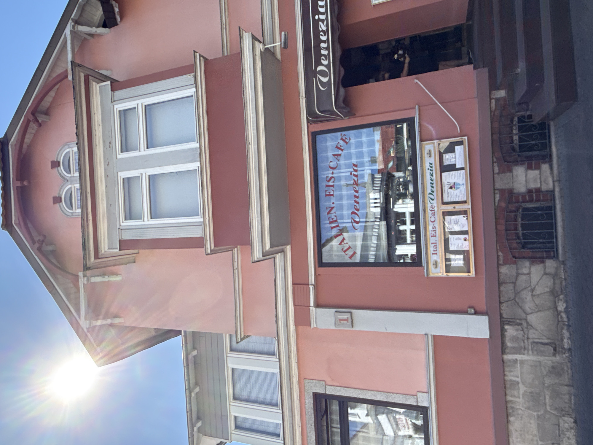 The image shows the exterior of an Italian ice cream cafe named "Ital. Eis-Café Venezia" located at Schulstraße 1 in Bad Sachsa, Germany. The building is painted in a salmon pink color with white and red trim. The cafe has large windows displaying the interior and the name of the establishment. Above one of the windows, a sign with the name "Venezia" is visible. To the right of the entrance, a person wearing a dark shirt and shorts is partially visible. The facade of the building features architectural details such as decorative brackets and a bay window. The ground level has a stone foundation with barred windows. The sun is shining brightly in the upper left corner of the image.