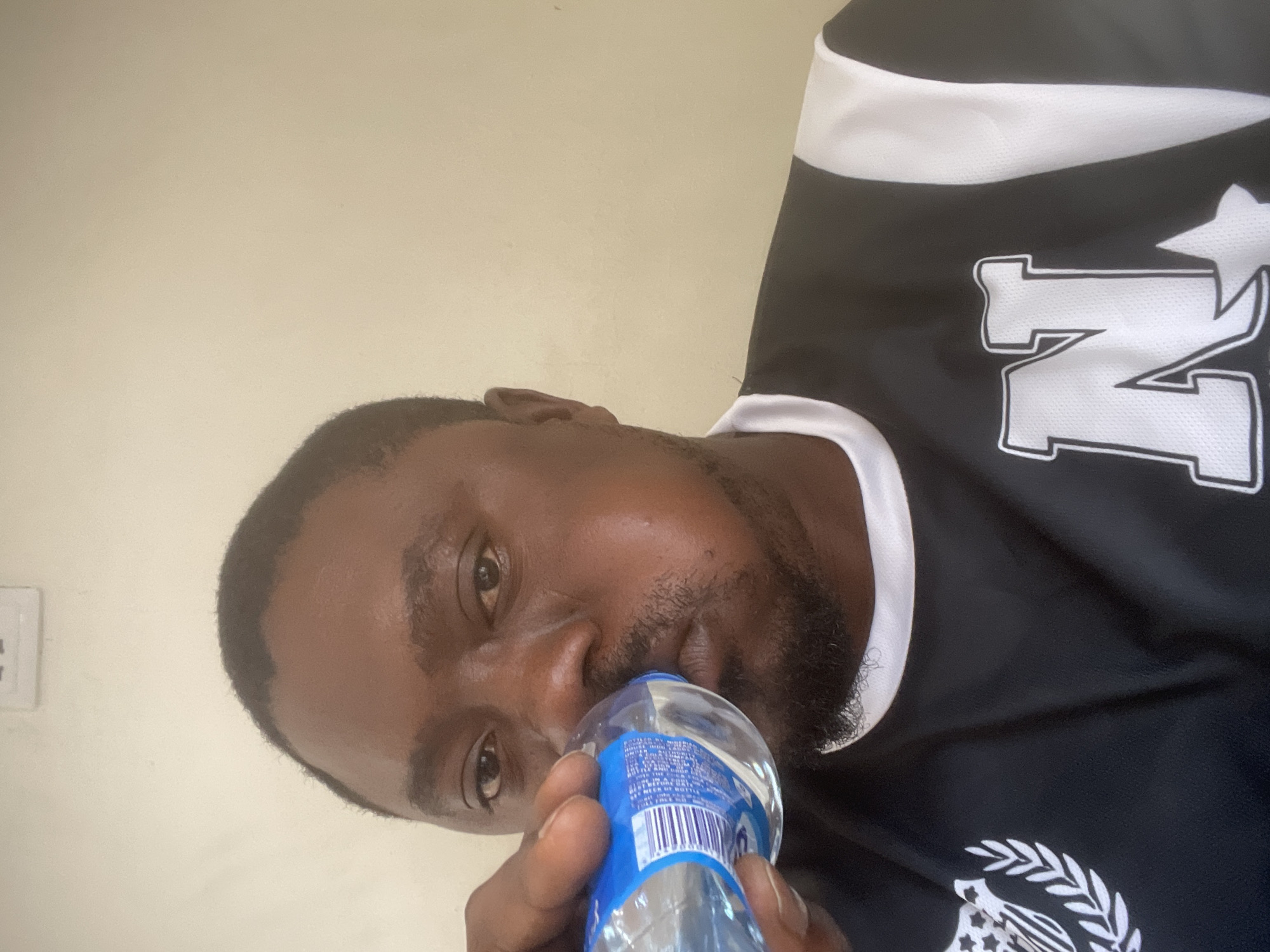 An adult man with dark skin, short dark hair, and a short beard is depicted in a close-up, portrait-style image, rotated 90 degrees counter-clockwise. He is looking directly at the camera with a neutral expression.
The man is holding a clear plastic water bottle with a blue label to his mouth, actively drinking or preparing to drink from it. He is wearing a black short-sleeved jersey or shirt, accented with white. On the right side of his chest/shoulder, a large white letter "N" with a white outline is visible. The lower left side of his shirt features a white emblem that includes a star, stripes, and laurel branches. White trim is also visible around the collar and sleeves.
The background consists of a plain, light-colored wall, appearing to be off-white or cream. A white electrical outlet or switch is partially visible on the wall to the left of the man's head. The lighting is even, suggesting an indoor setting. Small text on the water bottle's blue label is present but illegible, though a barcode is discernible. The image captures a moment of hydration, potentially in a personal or casual indoor setting in Jos, Nigeria.