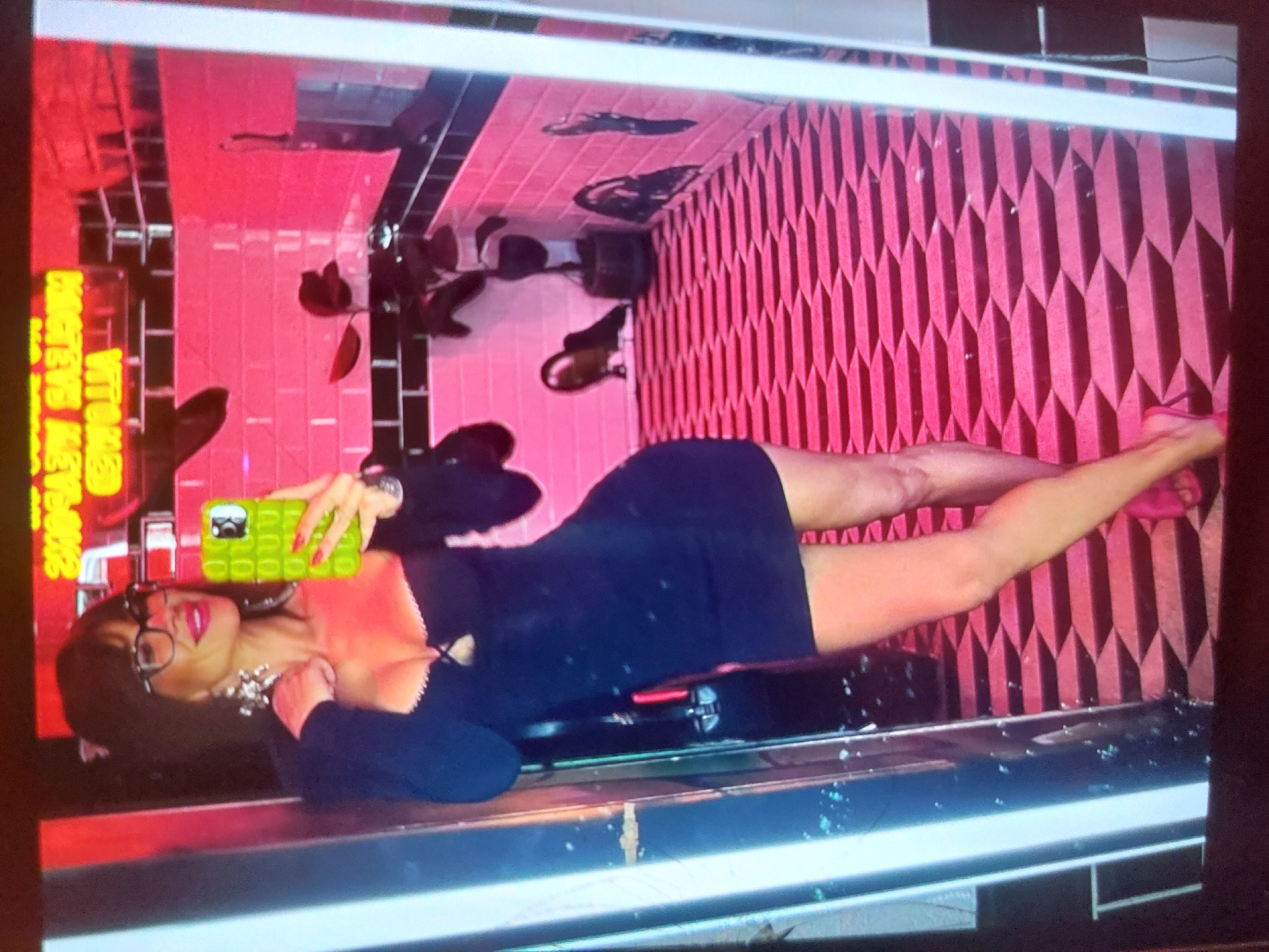 The image, when viewed upright, depicts a woman posing in a vibrant, pink and black-themed indoor setting, likely a bar or lounge in Tsarevo, Bulgaria. She is lying on a reflective, dark surface, extending her body towards the viewer. The woman has dark hair, wears glasses, red lipstick, a chunky silver necklace, and a long-sleeved, black mini-dress. Her legs are stretched out, with one foot clearly visible in a black high-heeled shoe. She holds a smartphone with a distinctive bright green pop-it case up towards her face, appearing to take a selfie or be captured in a photo.
The background features striking decor. One wall is covered in square pink tiles with black grout, while an adjacent wall boasts a textured, geometric pattern of pink diamond-shaped panels. Black square tiles are also integrated into the design. Several dark-leafed potted plants are visible, adding to the stylish ambiance. In the upper left area, a yellow neon sign is partially visible, displaying text that reads 'VITAMINS' or a similar word, and possibly 'PRETEVE' or 'PROTEIN' below it. The reflective surface she is on creates some visual interest with reflections of the surroundings. The overall scene conveys a sense of trendy leisure and a confident pose.