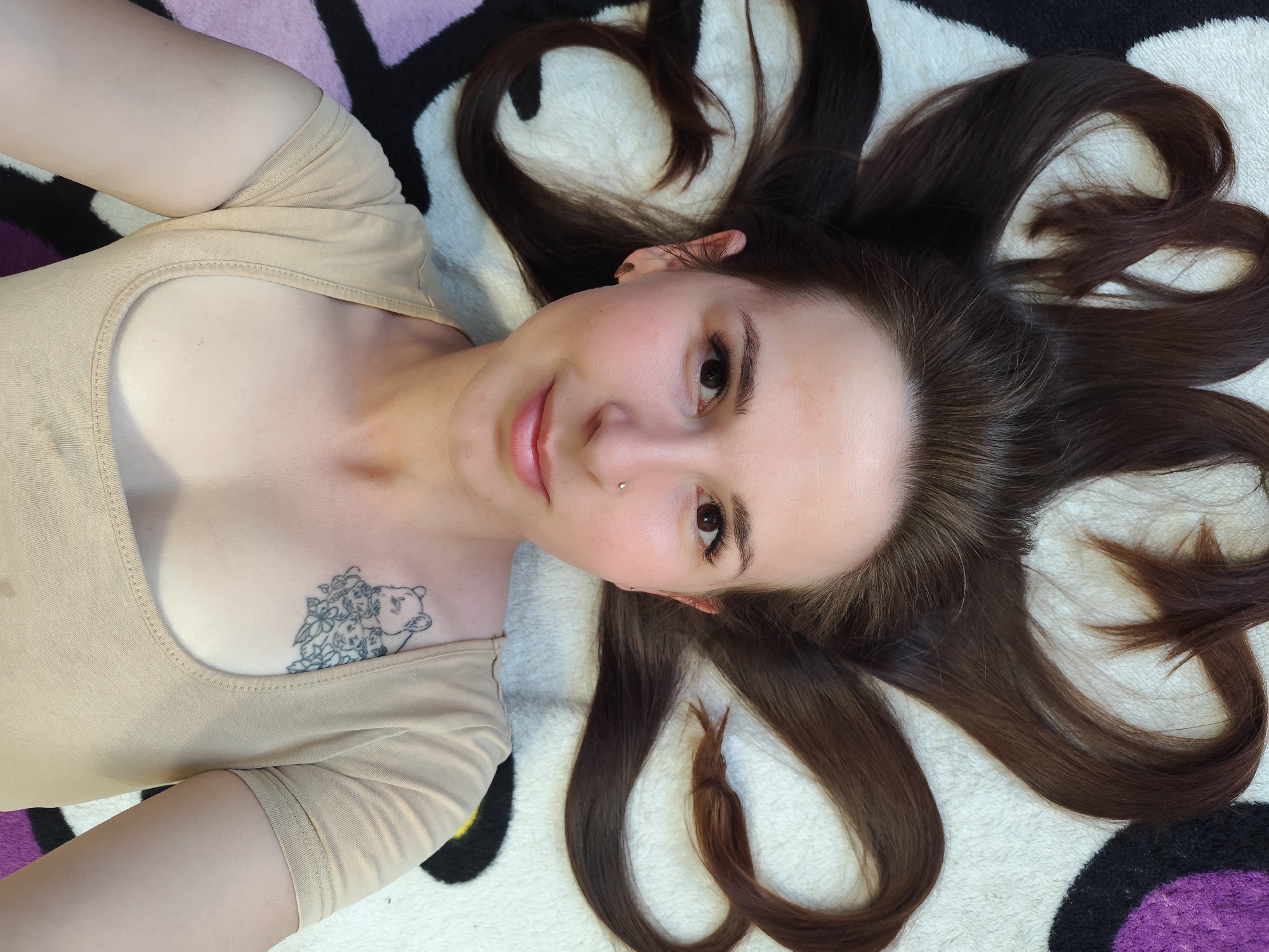 A young woman with brown hair lies on a patterned surface of black, white, and purple. She wears a beige scoop-neck shirt. A tattoo is visible on her chest. The image does not provide any information about a location in the city of Фоки, Russia.