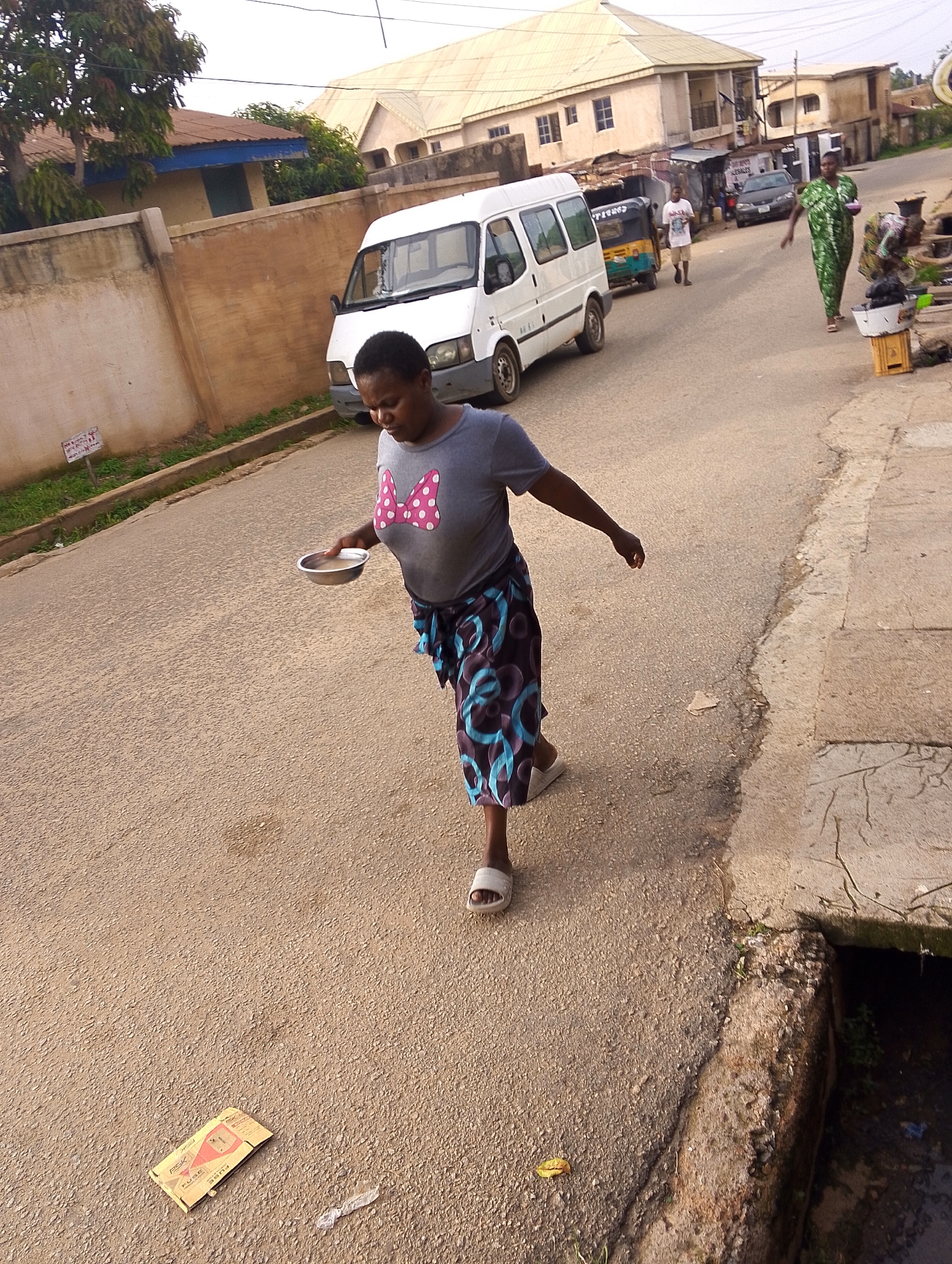 A woman in a gray t-shirt with a pink bow graphic and a patterned skirt walks on a street in Jos, Nigeria, holding a silver bowl of rice. She is wearing white sandals. In the background, a white van is parked on the street. Another woman in a green patterned dress walks in the background carrying a bowl and a baby is on her back. A third person, a man, walks away from the camera in the middle ground. Buildings line the street and the sky is overcast.