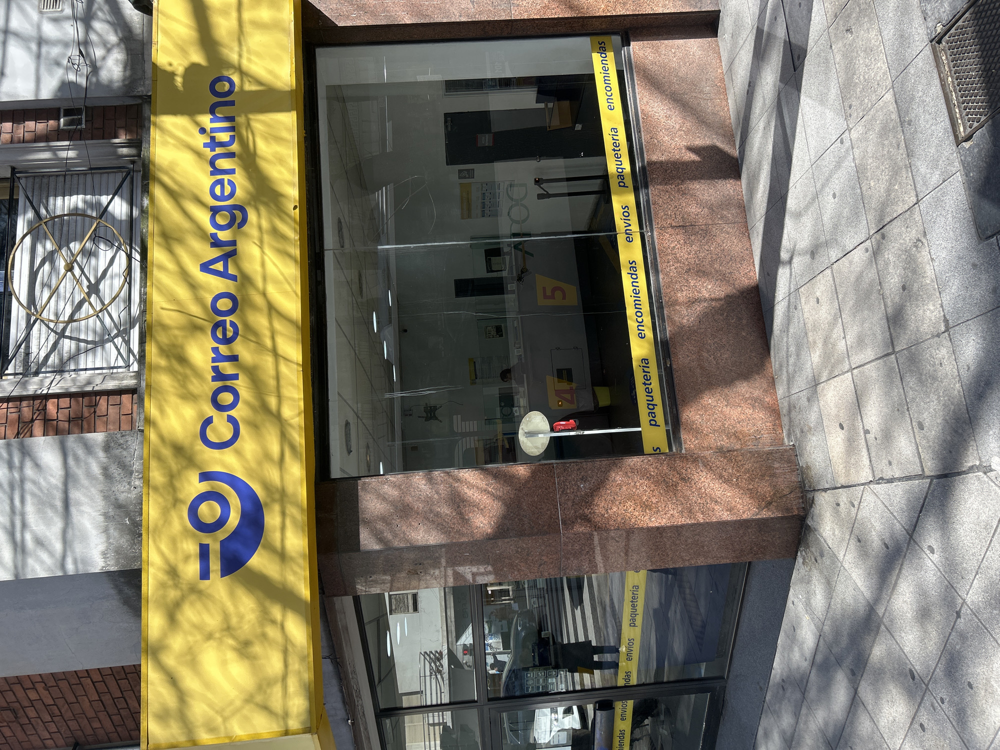 The image shows the exterior of a Correo Argentino post office, located in Buenos Aires, Argentina. The facade features a bright yellow awning with the blue Correo Argentino logo and text. Below the awning is a large glass storefront, revealing the interior of the post office. Inside, service counters and signage are visible. A yellow tape with the words "encomiendas envíos paquetería" is affixed to the lower part of the glass. The sidewalk in front of the building is made of gray paving stones and is partially covered in shadows cast by trees. The overall scene depicts a typical urban streetscape in a commercial area.