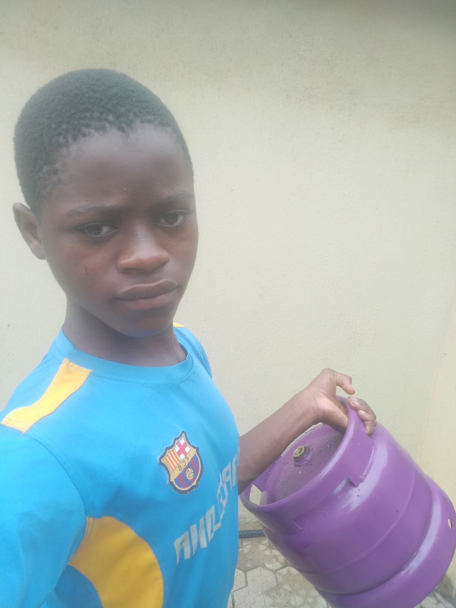 A young person wearing a blue and yellow soccer jersey with a Barcelona FC logo is holding a purple gas cylinder. The background is a light beige wall and possibly a paved ground surface. The location is the city of Shangisha, Nigeria.
