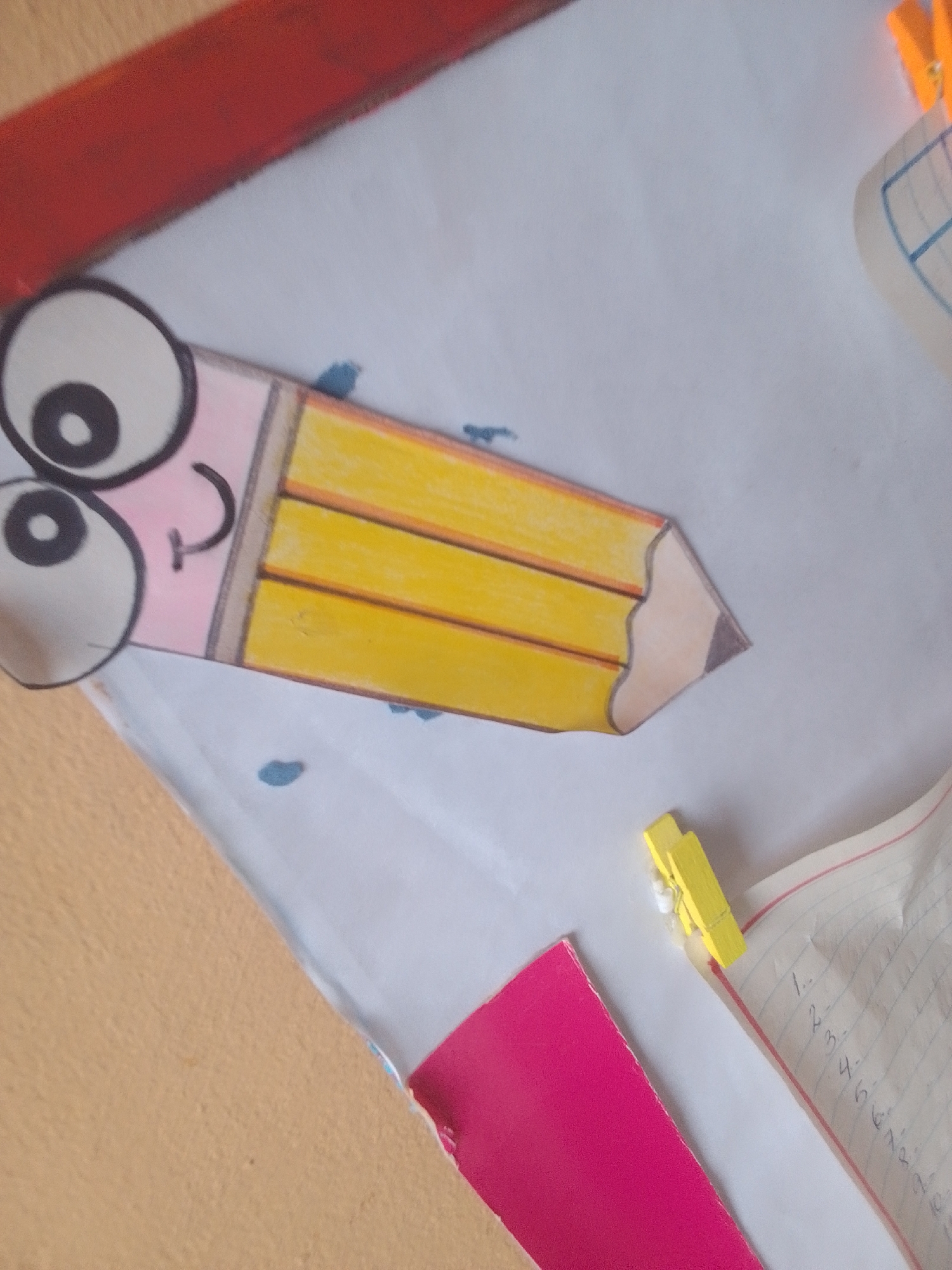 The image shows a cartoon drawing of a pencil with a face, placed on a white surface. The pencil has large, round eyes with black pupils and a smiling mouth. Its body is yellow with orange stripes, and the tip is sharpened and colored in shades of brown and tan. The drawing appears to be cut out from paper.
To the left of the drawing, a portion of a light brown surface is visible, possibly a table or desk. Above the drawing, a red object, possibly a ruler or border, runs horizontally across the top of the frame. In the background, there are small blue shapes, possibly ink blots or part of another drawing.
To the right of the pencil drawing, two yellow clothespins are clipped onto a white paper with red lines, resembling a notebook or practice sheet. Numbers are written on the paper, appearing to be a list, starting with "1." and continuing with subsequent numbers. A pink piece of paper is also visible in the lower right quadrant, partially obscuring the notebook. The lighting suggests an indoor setting, and the overall scene appears to be related to schoolwork or a craft activity. There is no indication of time of day, weather, or specific emotions beyond the cartoon's cheerful expression. The location context provided is Santa Ana, Venezuela.