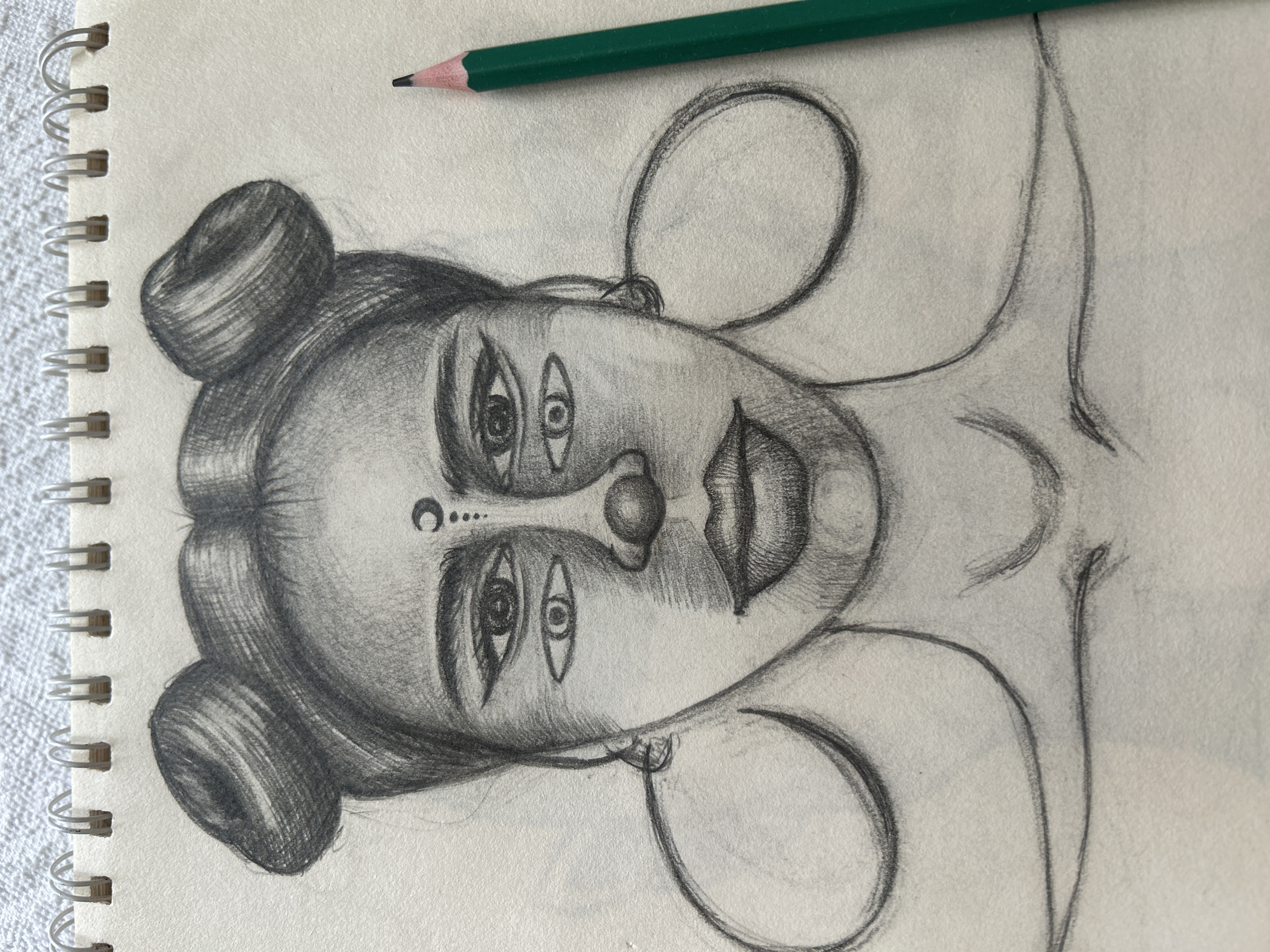 A pencil sketch of a woman's face with multiple eyes and a crescent moon symbol on her forehead is shown in a spiral-bound notebook. The drawing is done in grayscale and features a green pencil lying to the right of the notebook. The background appears to be a textured surface, possibly a fabric. There is no indication of a specific location in Moscow, Russia, or any actions or interactions occurring.