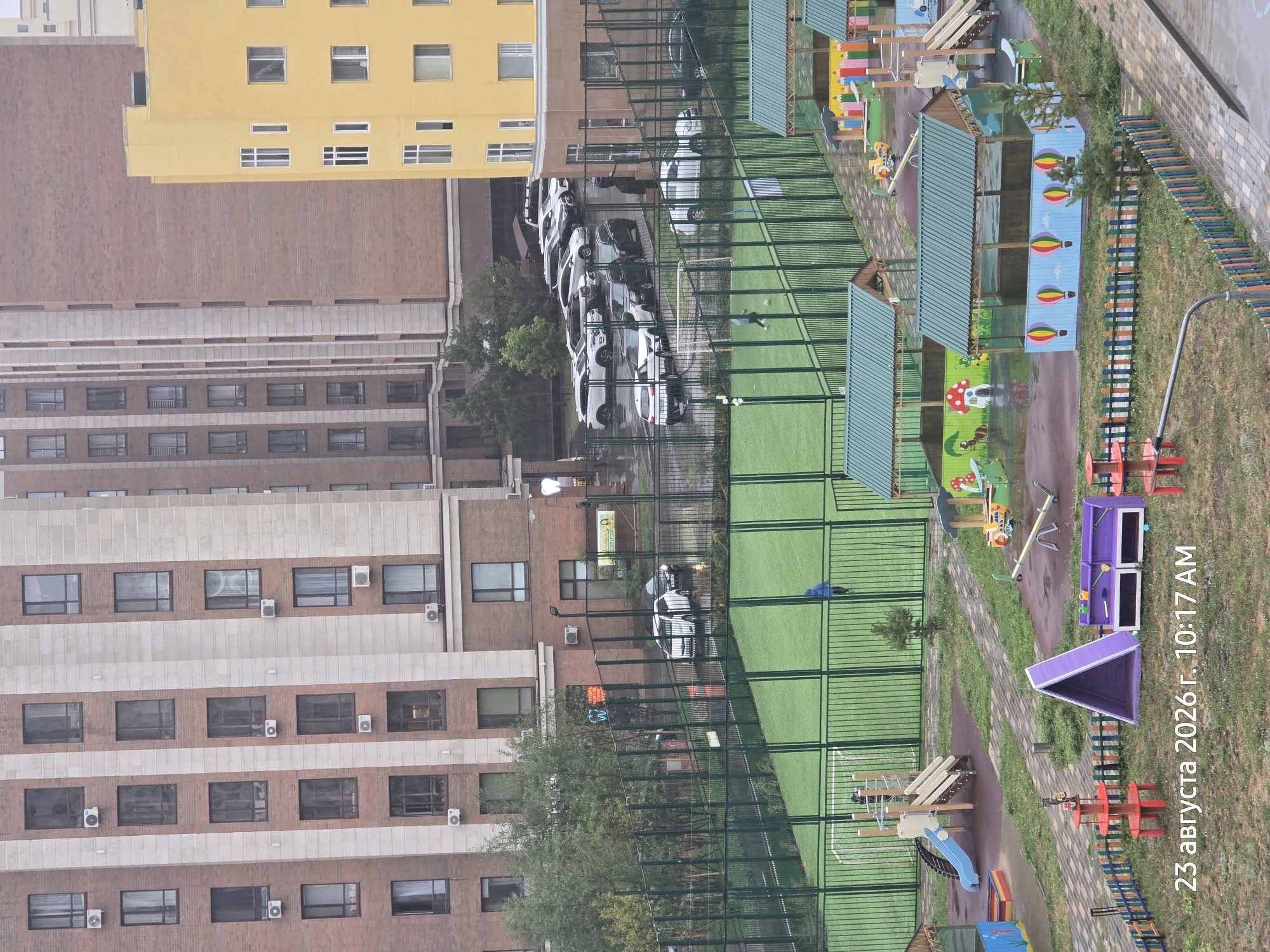 In Astana, Kazakhstan, a residential complex features multi-story buildings with brick and light-colored facade elements. A yellow building stands to the right. In front of the buildings, there's a fenced-in area with a green artificial turf sports field, possibly for soccer, with a goal at one end. A single person in a blue jacket is visible on the field. Beyond the sports field is a playground with various colorful structures, including slides, swings, a seesaw, and covered playhouses with decorative paintings. Several cars are parked in an adjacent lot behind the fence. The ground appears wet, suggesting recent rain. The overall scene depicts a modern urban residential area with recreational facilities.