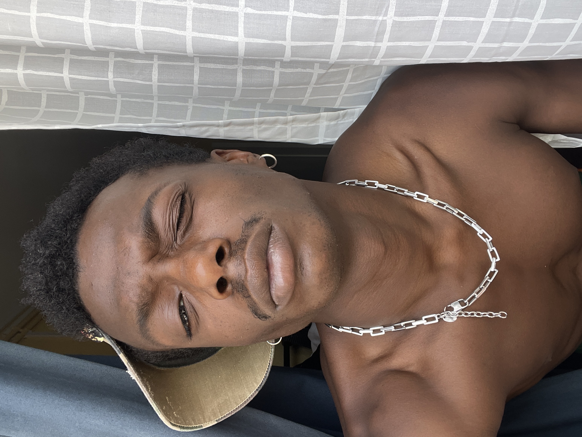 A man is posing for a selfie, half-naked, wearing a silver chain and a cap. He is looking at the camera and winking. The background shows a dark curtain on the left and a white patterned curtain on the right. The lighting suggests an indoor setting, possibly near a window. The scene is an urban environment, but no specific location within Barcelona is identifiable.