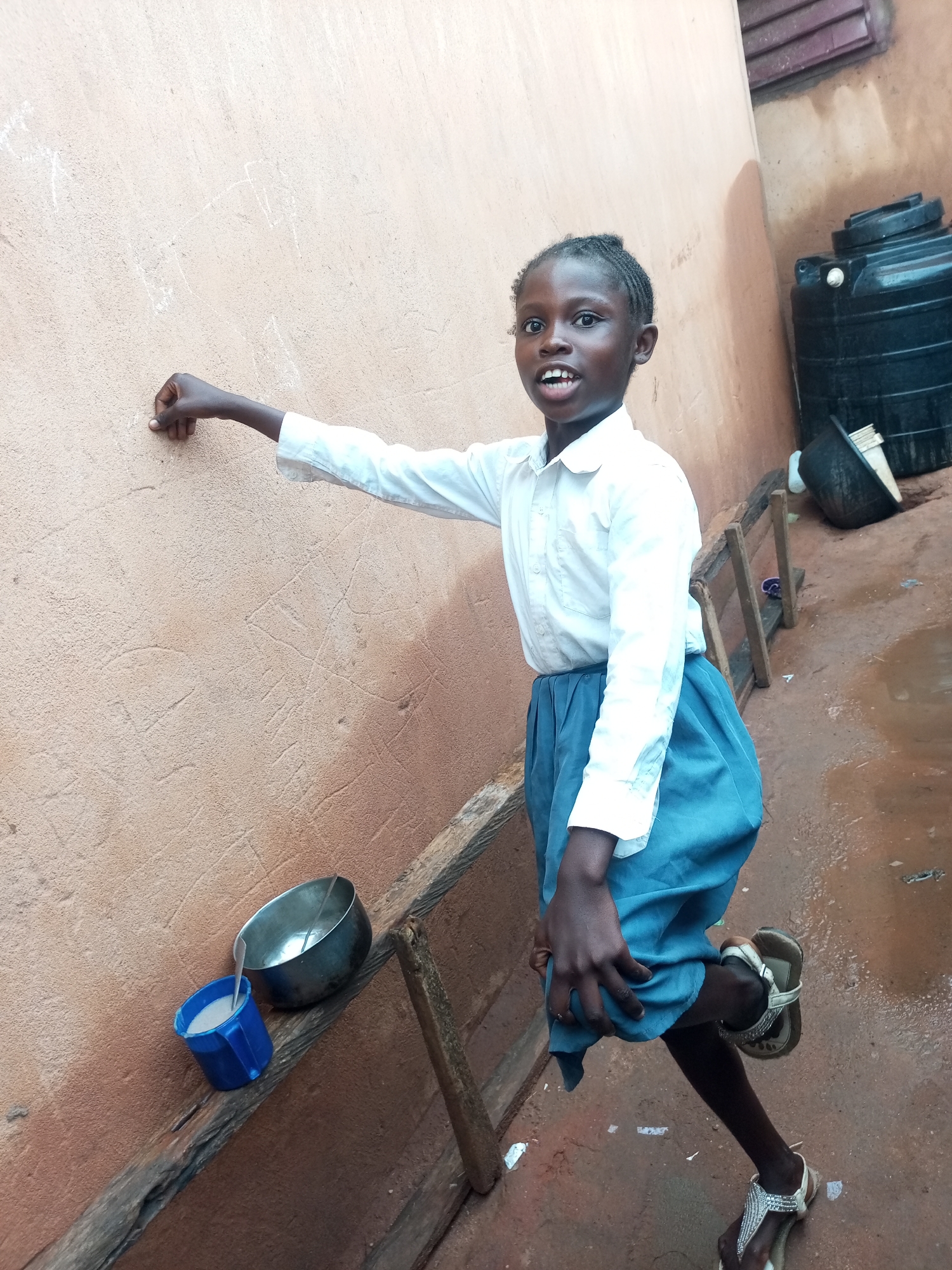 A young Black girl, wearing a white collared shirt and a blue skirt, stands outdoors. She is facing slightly to the right, looking at the camera with her mouth open. Her left arm is extended, and her index finger is touching a light brown, textured wall. There are some faint white markings on the wall, possibly chalk drawings or scratches.
In the lower left of the image, on a wooden beam, are two containers: a blue plastic mug filled with a white liquid and a larger metal bowl. To the right of the girl, a large black water tank is visible against a darker, red-brown wall. The ground appears to be wet and muddy, with some scattered debris. The overall environment suggests an outdoor area in a residential or communal space.
This scene is located in the city of Tempure, Nigeria.
