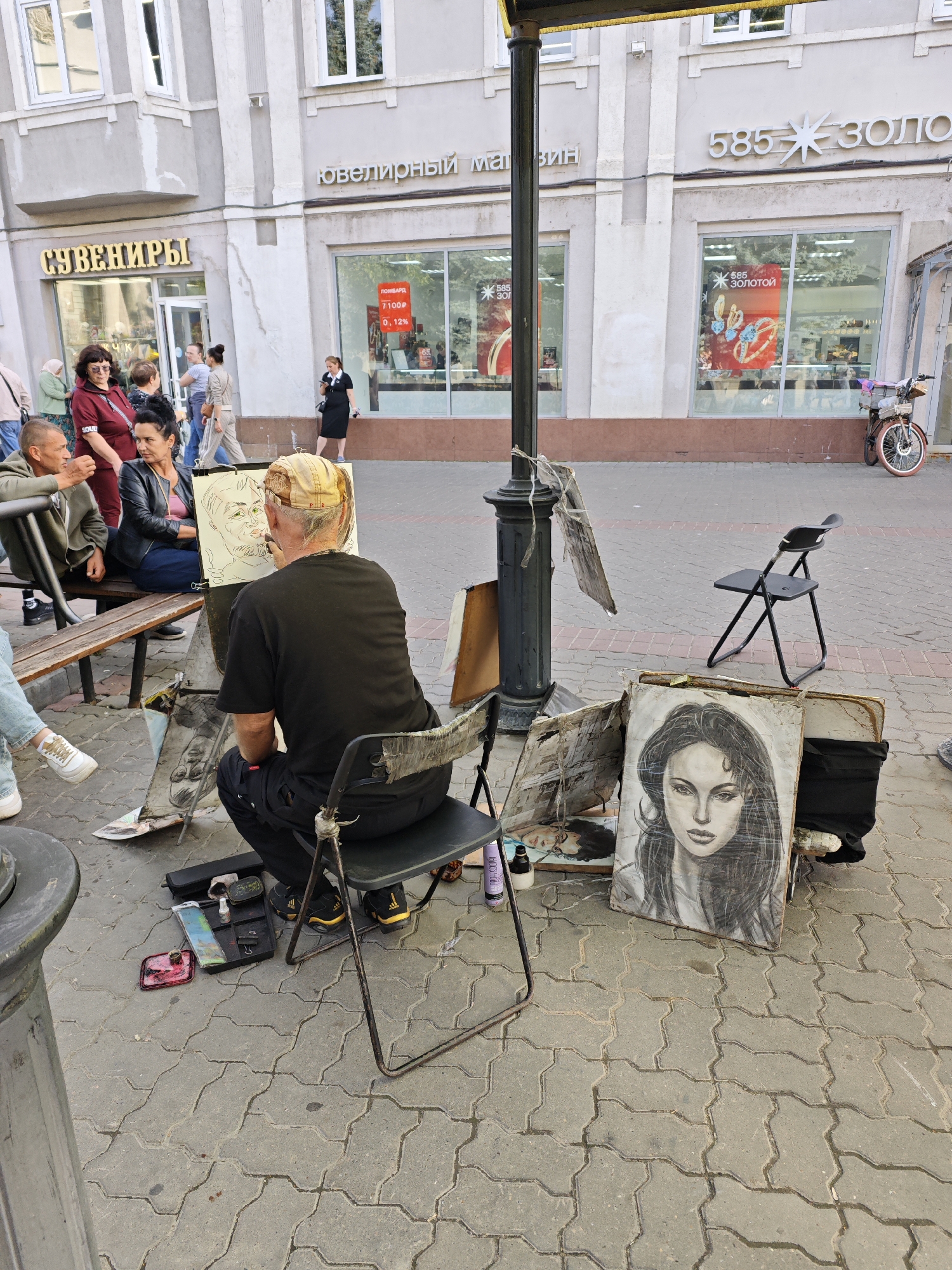 An outdoor scene shows a man drawing a portrait on the sidewalk in Kazan, Russia. The man, wearing a black t-shirt and a backward baseball cap, is seated on a folding chair, facing an easel displaying a sketch of another person. Next to the easel, a finished portrait of a woman with long dark hair is propped up. Various art supplies, including brushes, pencils, and paints, are scattered on the ground.
Several people are visible in the background, some observing the artist. A bicycle is parked to the right. The setting appears to be a pedestrian area with shops lining the street. Signs above the shops read "СУВЕНИРЫ" (Souvenirs) and "585 ЗОЛОТО" (585 Gold), indicating the presence of retail stores. The weather appears to be overcast, and the time of day is likely afternoon. The overall mood is relaxed and artistic.