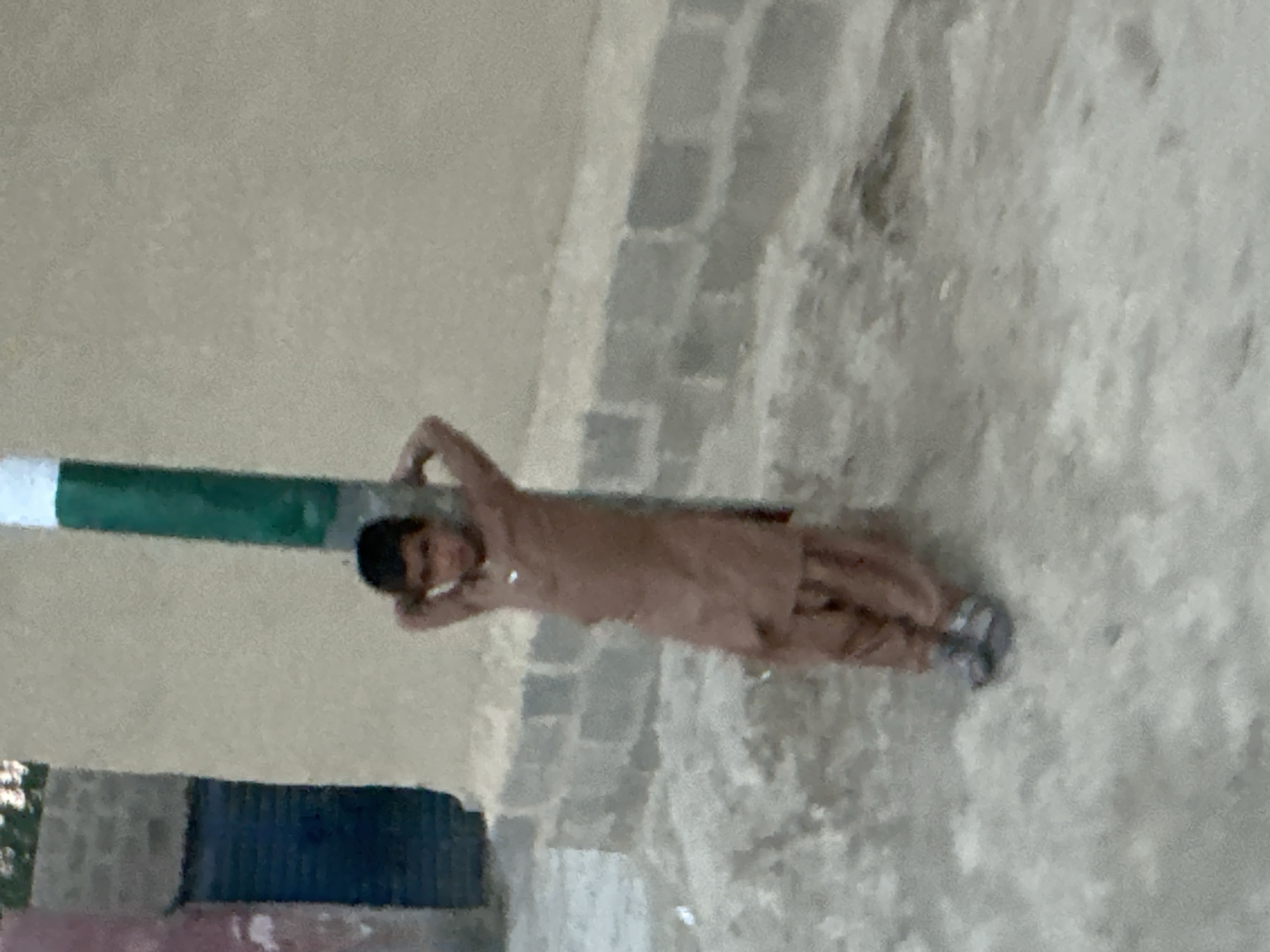 A blurry, low-resolution photograph, rotated sideways, depicts a young boy lying casually on the ground in an outdoor setting. The boy, with dark hair, is wearing a light brown tunic, possibly a shalwar kameez, and grey sandals. He is leaning on his side against a green and white striped pole or pillar, with his arms bent and hands near his head, adopting a relaxed pose. A slight smile or smirk is visible on his face.
He rests on dusty or sandy ground, adjacent to what appears to be a low wall or foundation constructed from grey bricks or blocks. In the background, there is a dark, possibly blue, rectangular structure or object, and vague hints of greenery. The lighting suggests it is daytime, but there are no strong shadows, possibly indicating an overcast sky. The overall scene conveys an informal and simple environment, likely a courtyard, street, or rural area. No text is visible in the image.