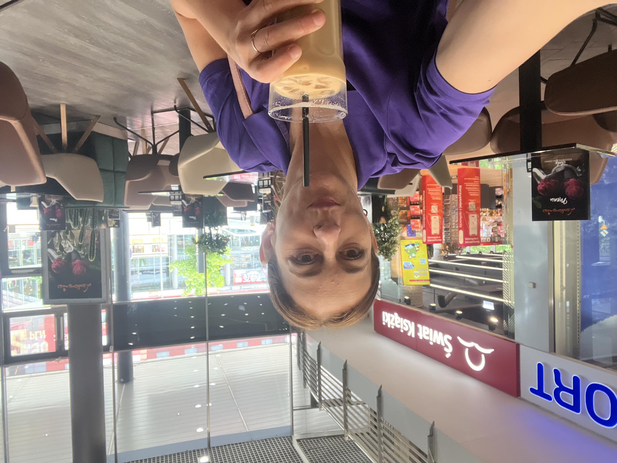 A woman with light brown hair is taking a selfie in a cafe, holding a glass of iced coffee. She is wearing a purple shirt and a gold ring on her right hand. In the background, there are tables and chairs, as well as shops with signs in Polish. One shop has a sign that says "Świat Książki," which translates to "World of Books." Another sign says "ORT." The cafe appears to be located in a mall or shopping center.