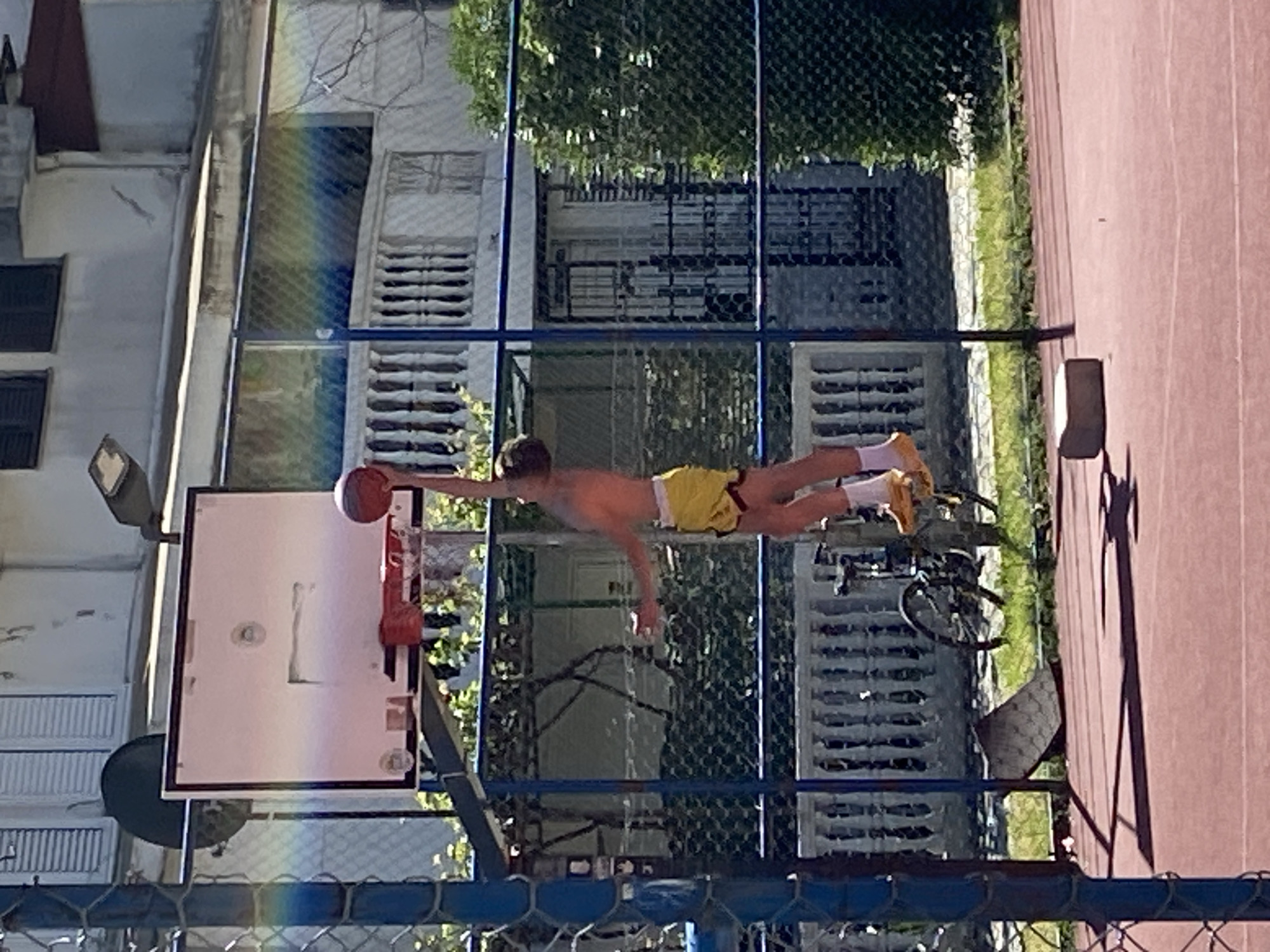 A basketball court in Budva, Montenegro, shows a shirtless individual in yellow shorts leaping towards a basketball hoop with a basketball in hand. A building with louvred windows is on the left, and a green fence is in the background. A bicycle is parked nearby. The ground is red. A streetlight is also visible.
