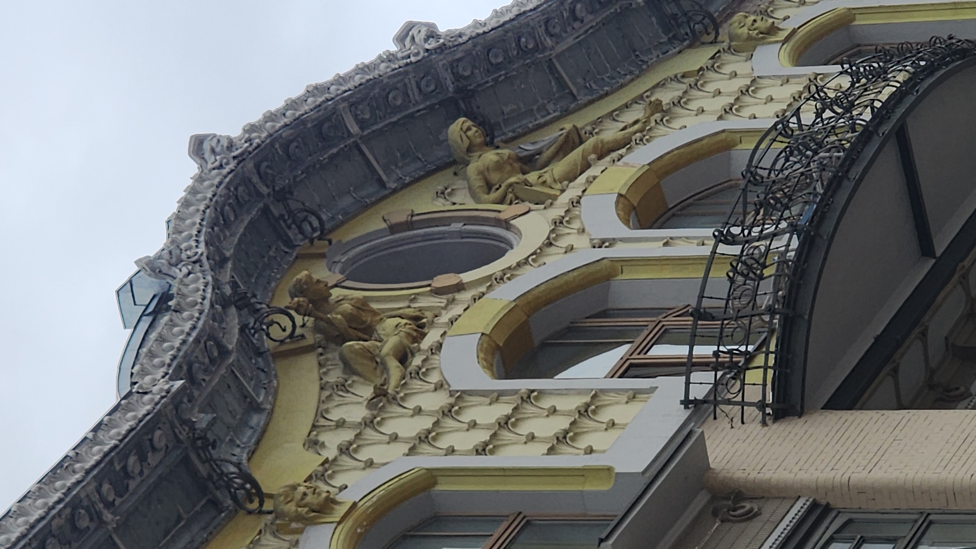 The image shows a close-up view of a building facade with intricate architectural details. The building features decorative moldings, sculptures, and ornate metalwork. The architectural style appears to be classical or baroque, characterized by the use of relief sculptures, arched windows, and a decorative balustrade. The sculptures include human figures in reclining poses, adding to the artistic value of the facade. The building's color scheme consists of a combination of light yellow, white, and gray elements, enhancing its visual appeal. The building might be located in the city of Moscow, Russia. The environment is cloudy or overcast, casting soft light on the building.