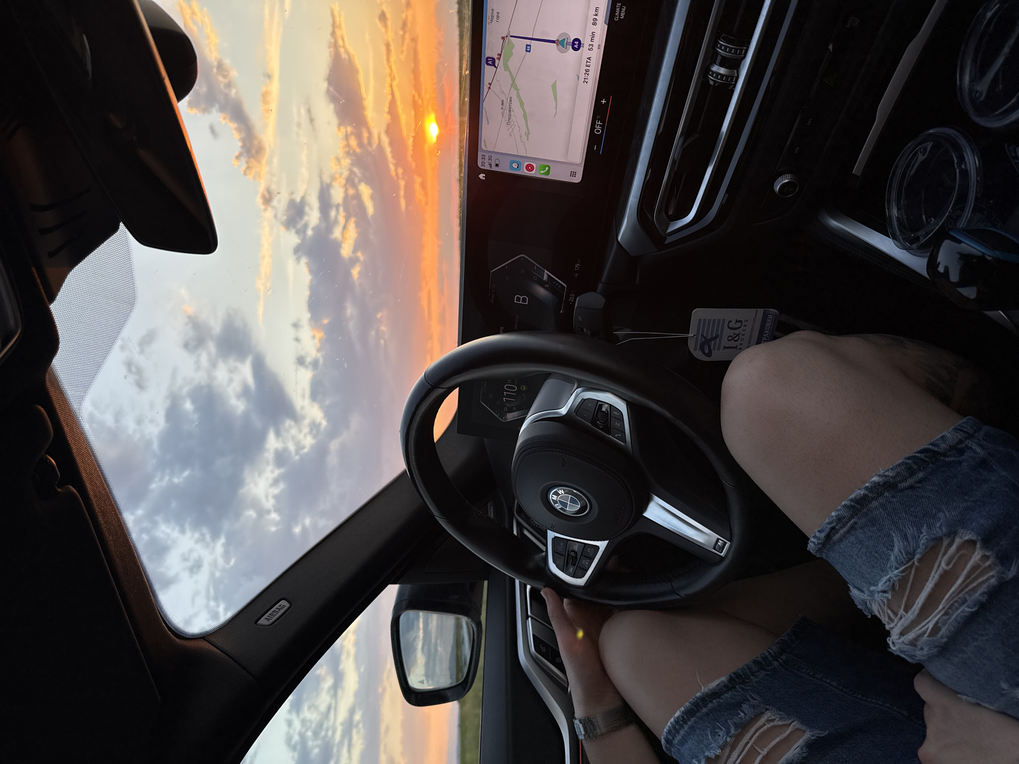 The photograph is taken from inside a car, capturing a view through the windshield toward a sunset. The dashboard, steering wheel, and center console are prominently visible, and the car's navigation screen displays a map with a route planned. The map indicates that the destination is 89 kilometers away and is near a place called Градина in Bulgaria. The estimated time of arrival (ETA) is 21:26. A person is seen wearing denim shorts, with the fabric torn near the knee. The steering wheel is held in one hand. The sky visible through the windshield shows a mix of clouds and the setting sun.